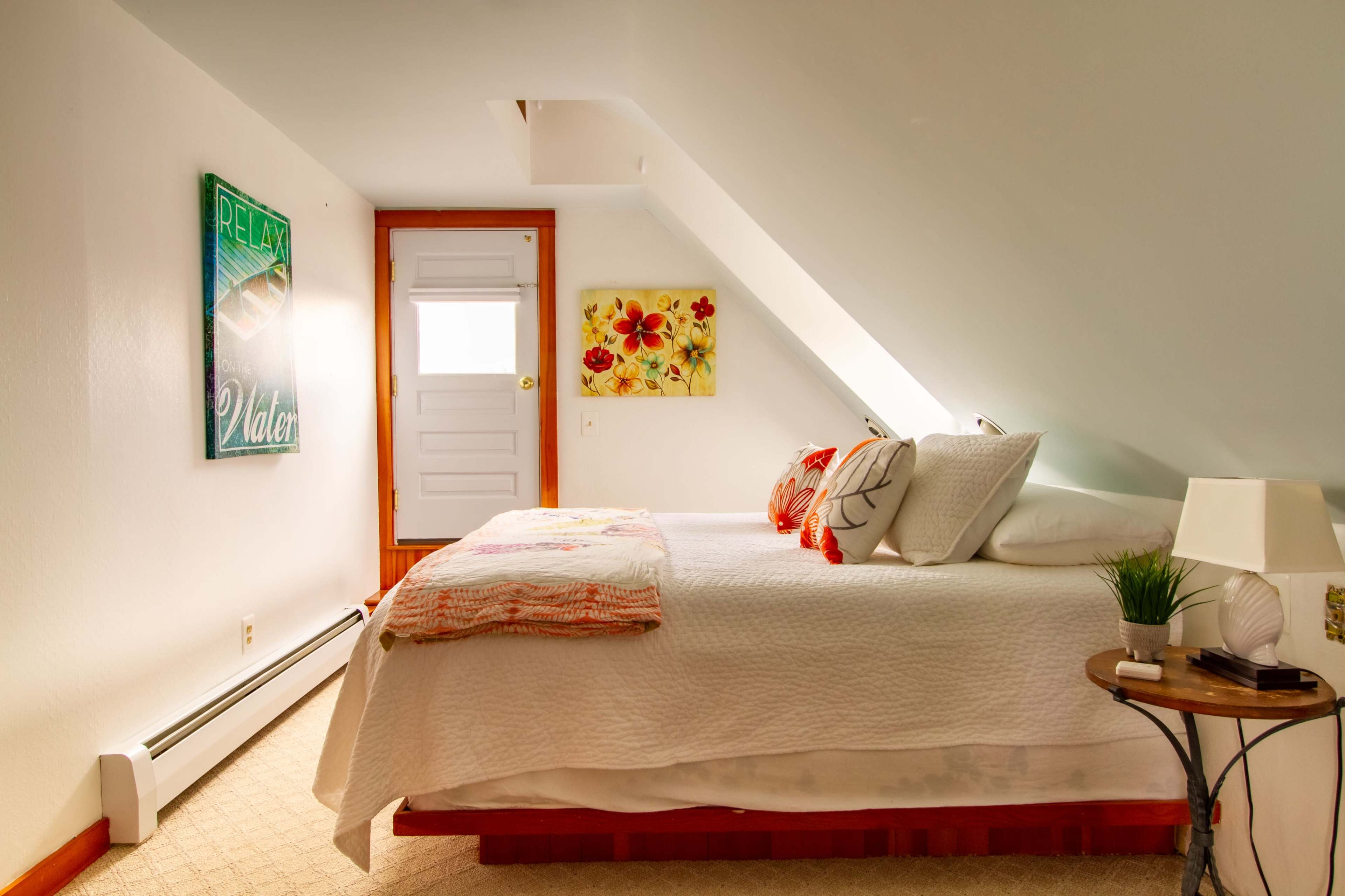 A neatly arranged bedroom features a double bed with patterned pillows, artwork on the walls, a small table beside the bed, and a door leading outside.