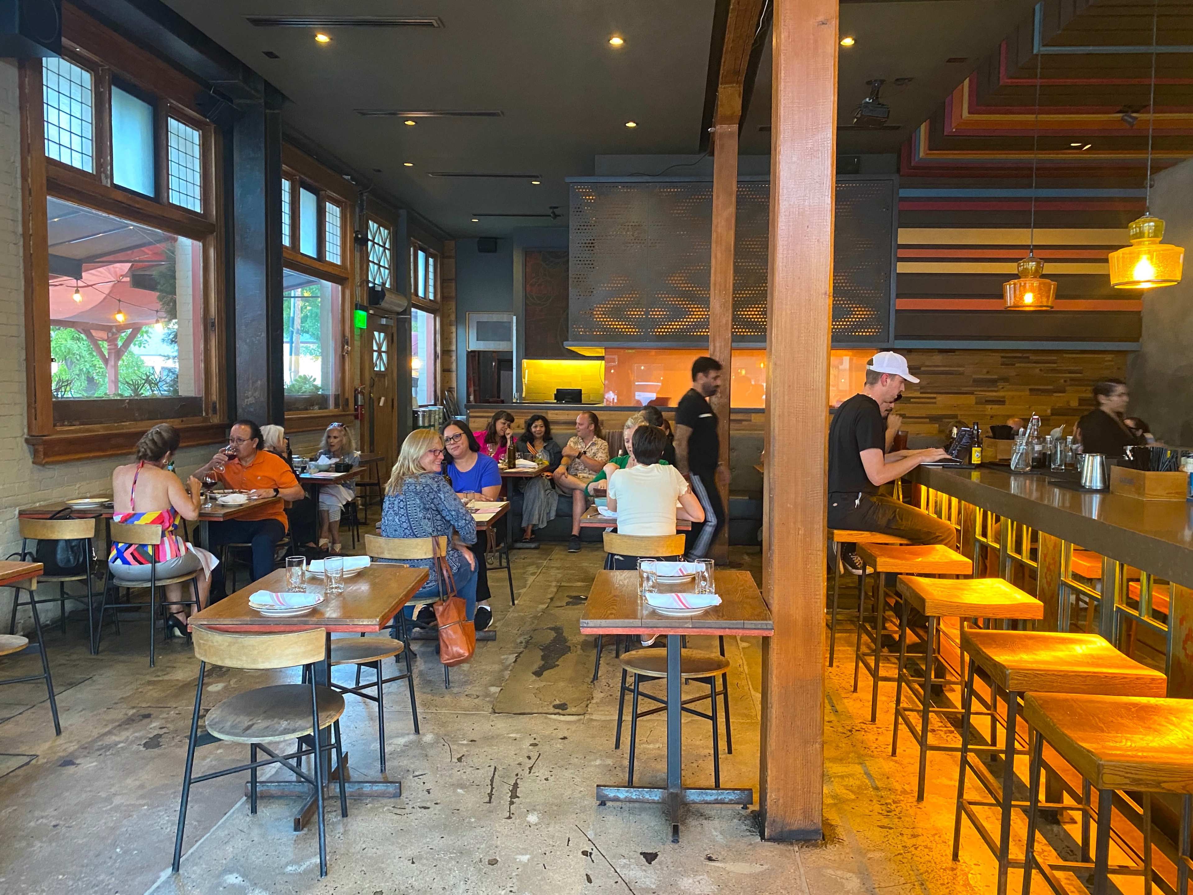 The interior of a restaurant shows several groups of patrons seated at tables and the bar, with staff moving between them.