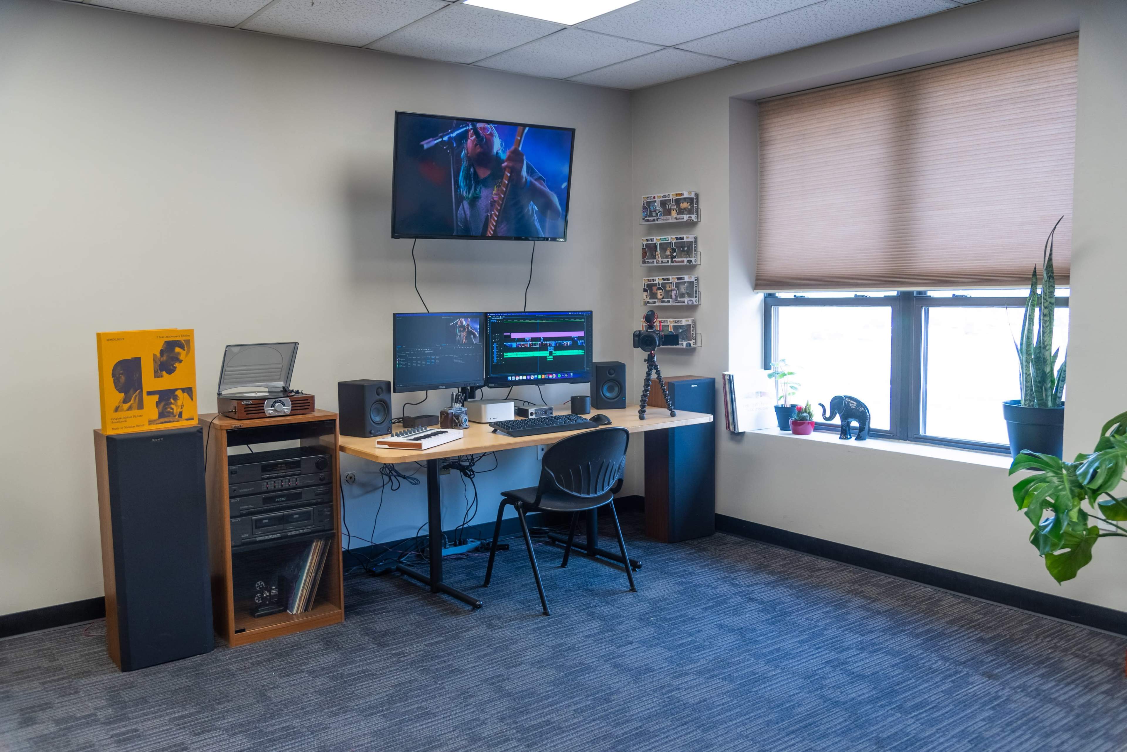 A home office or editing suite with a large desk, two computer monitors, audio speakers, and various equipment, including a camera and a plant near a window.