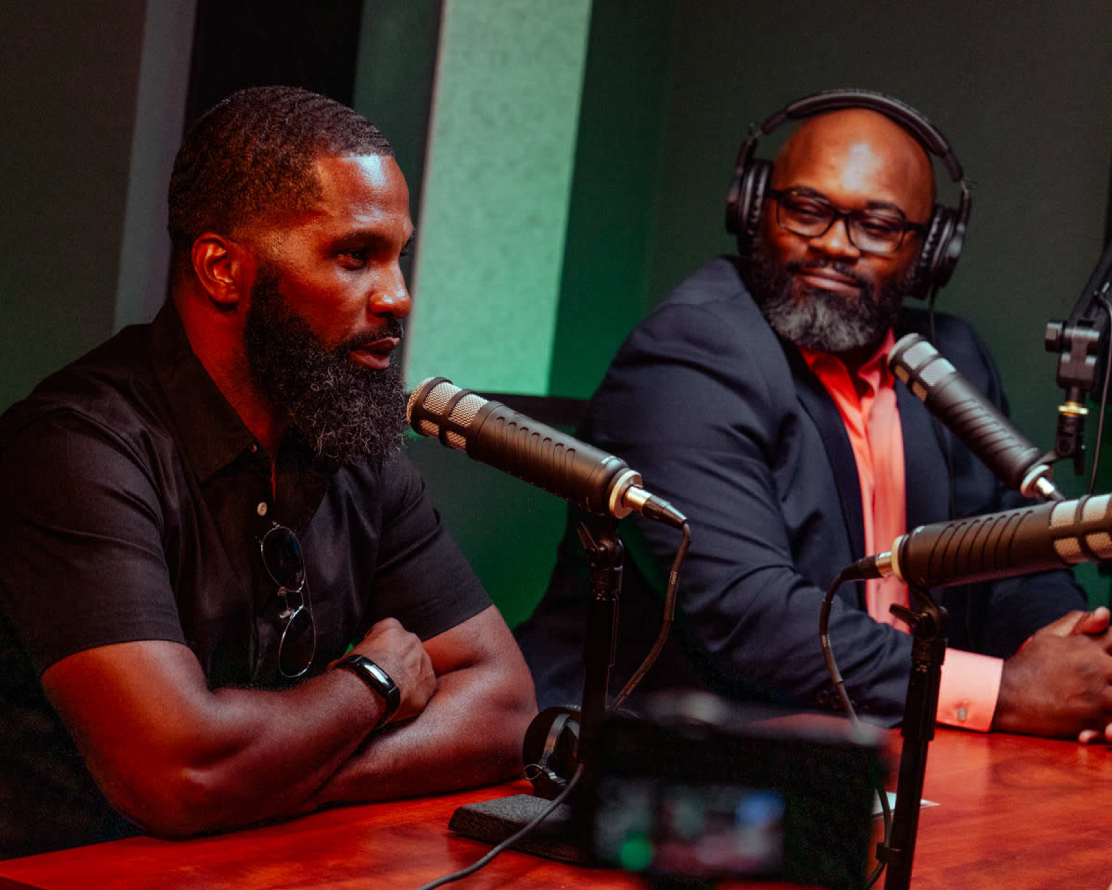 Two men are seated at a table with microphones in front of them, engaged in a discussion in a recording studio.