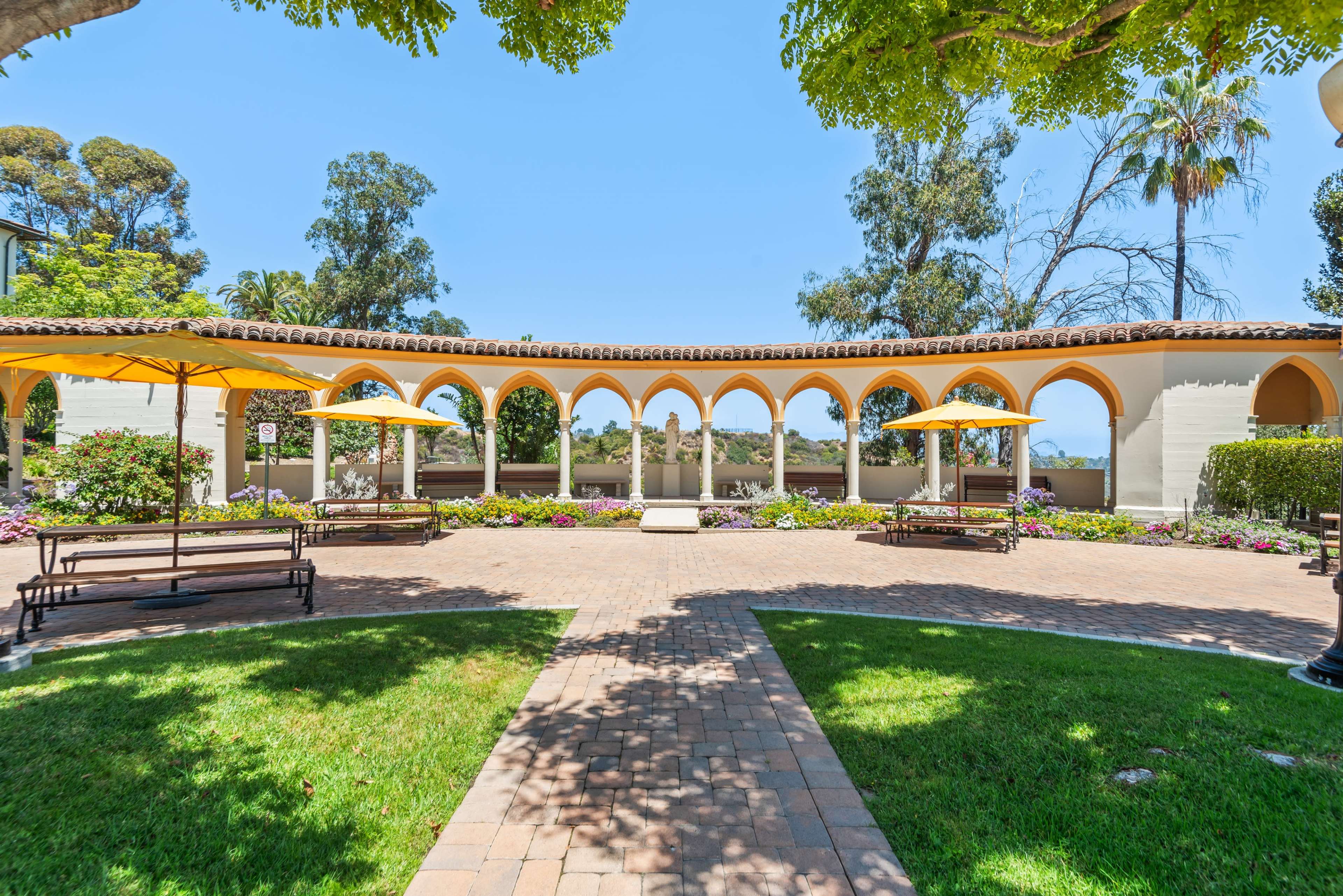 The image shows a landscaped courtyard with arched walkways, benches, and yellow umbrellas, surrounded by trees and flowering plants.