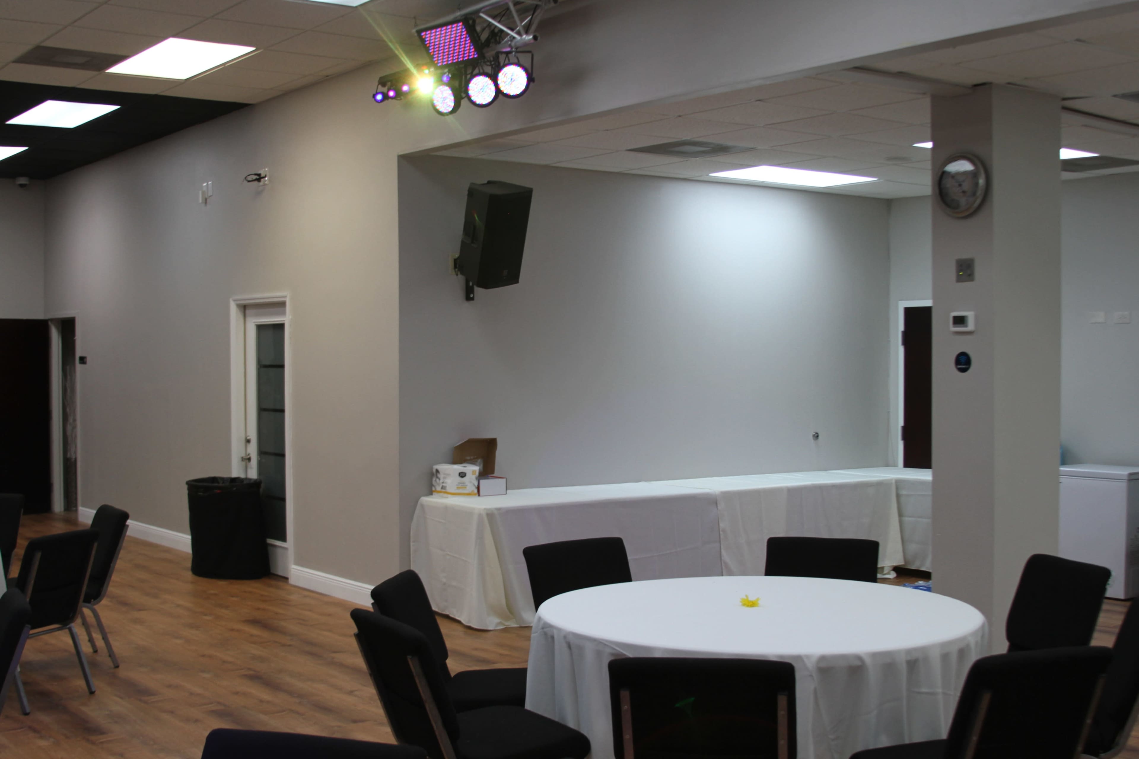The image shows a spacious indoor event space with a round table set up and a stage area featuring lighting and a speaker.