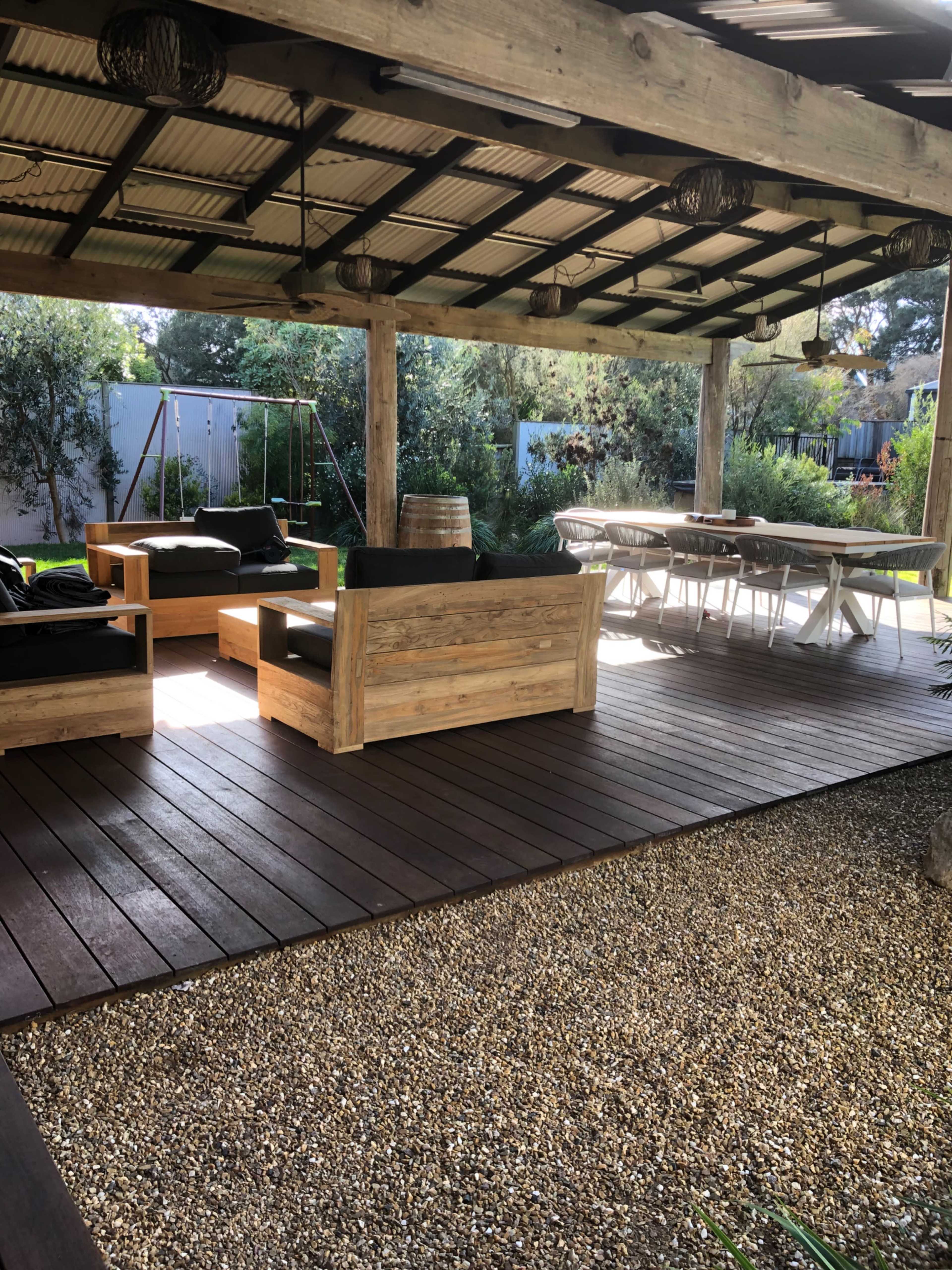 A shaded outdoor seating area with wooden furniture, a gravel floor, and a dining table under a roofed patio.