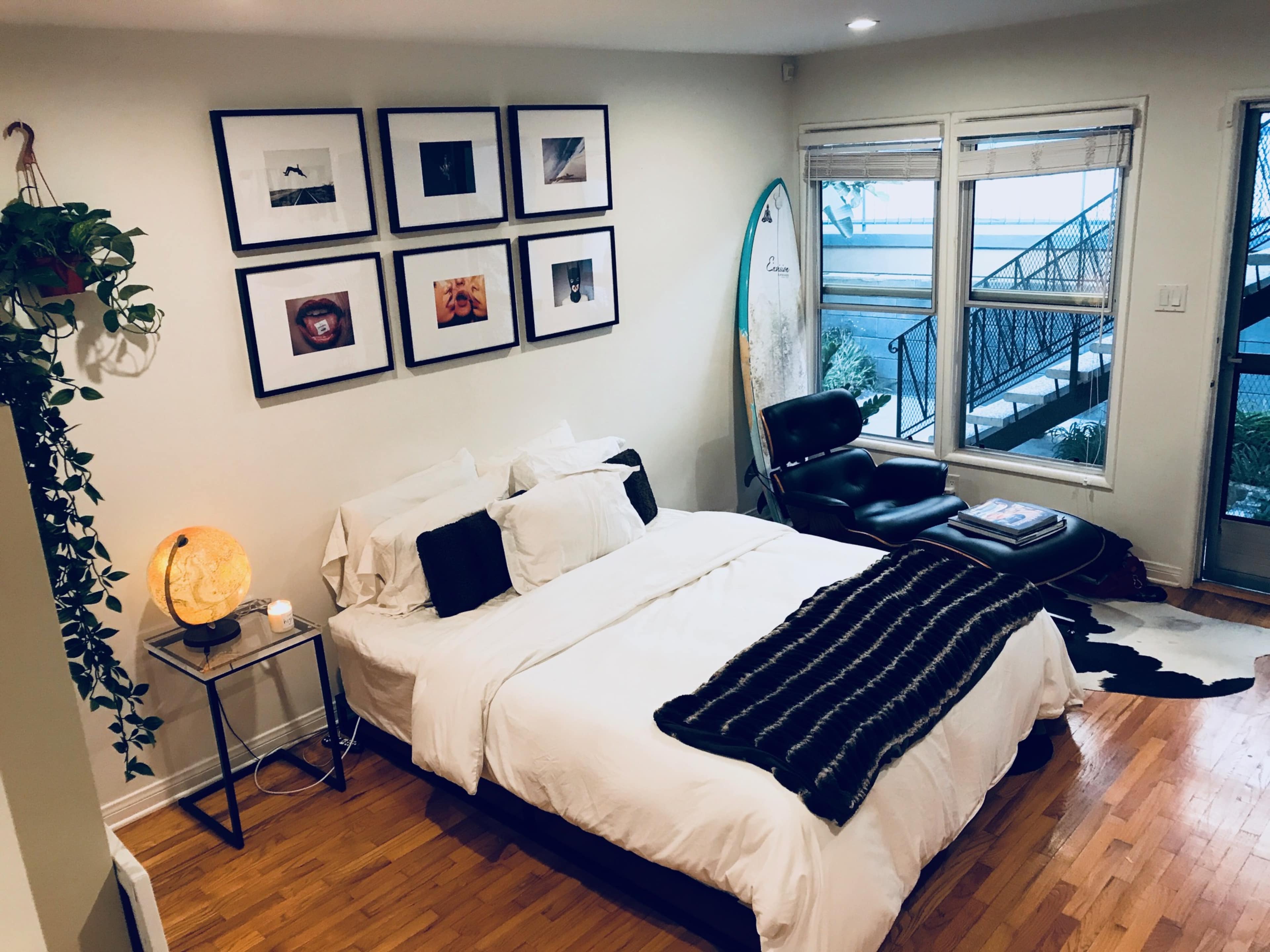 The room features a bed with white bedding and black accents, framed photos on the wall, a surfboard leaning against the wall, and a chair beside a window.