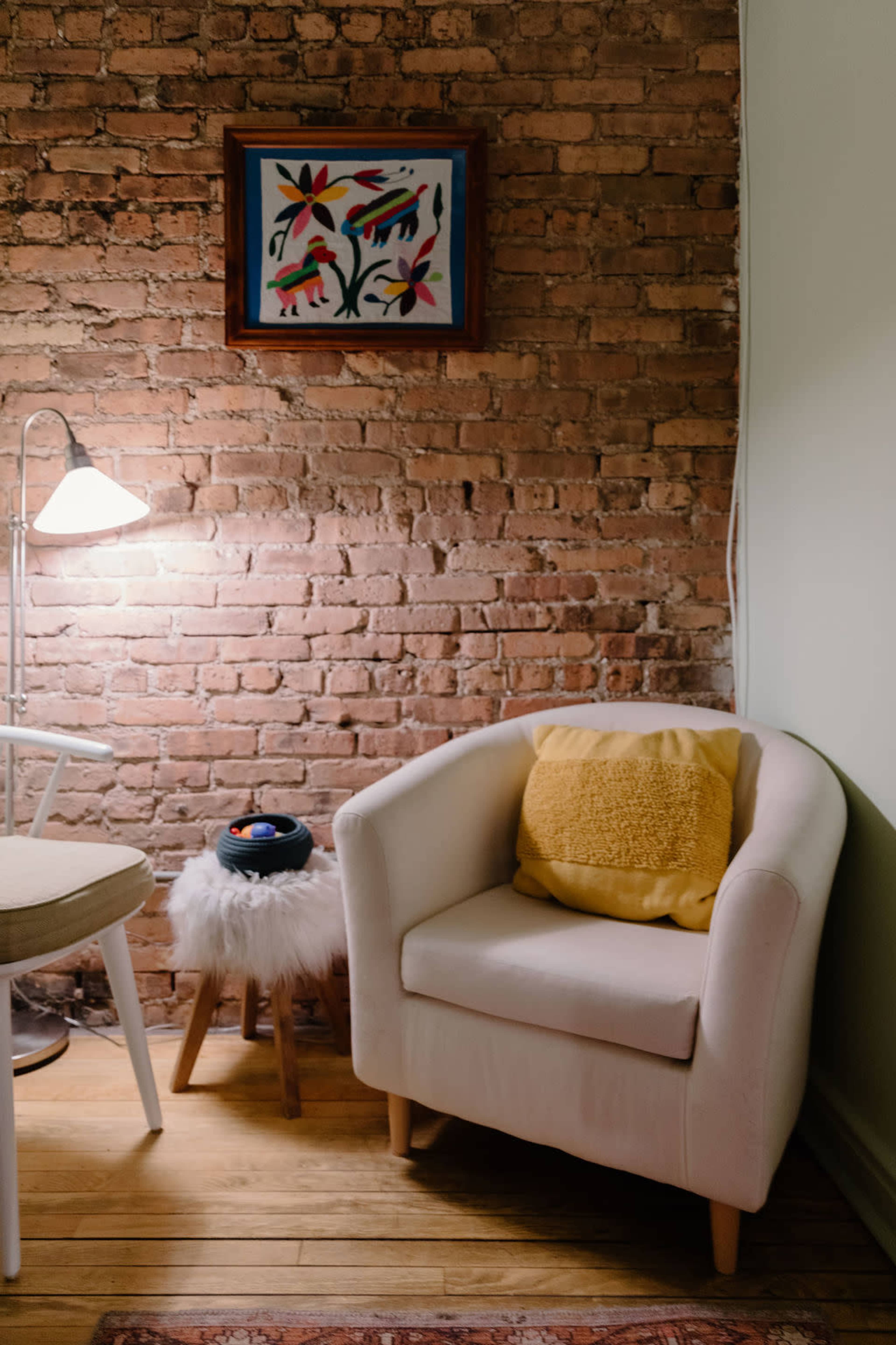 A cozy seating area features a white armchair with a yellow pillow, a small wooden stool topped with decorative items, and a floor lamp beside a textured brick wall.