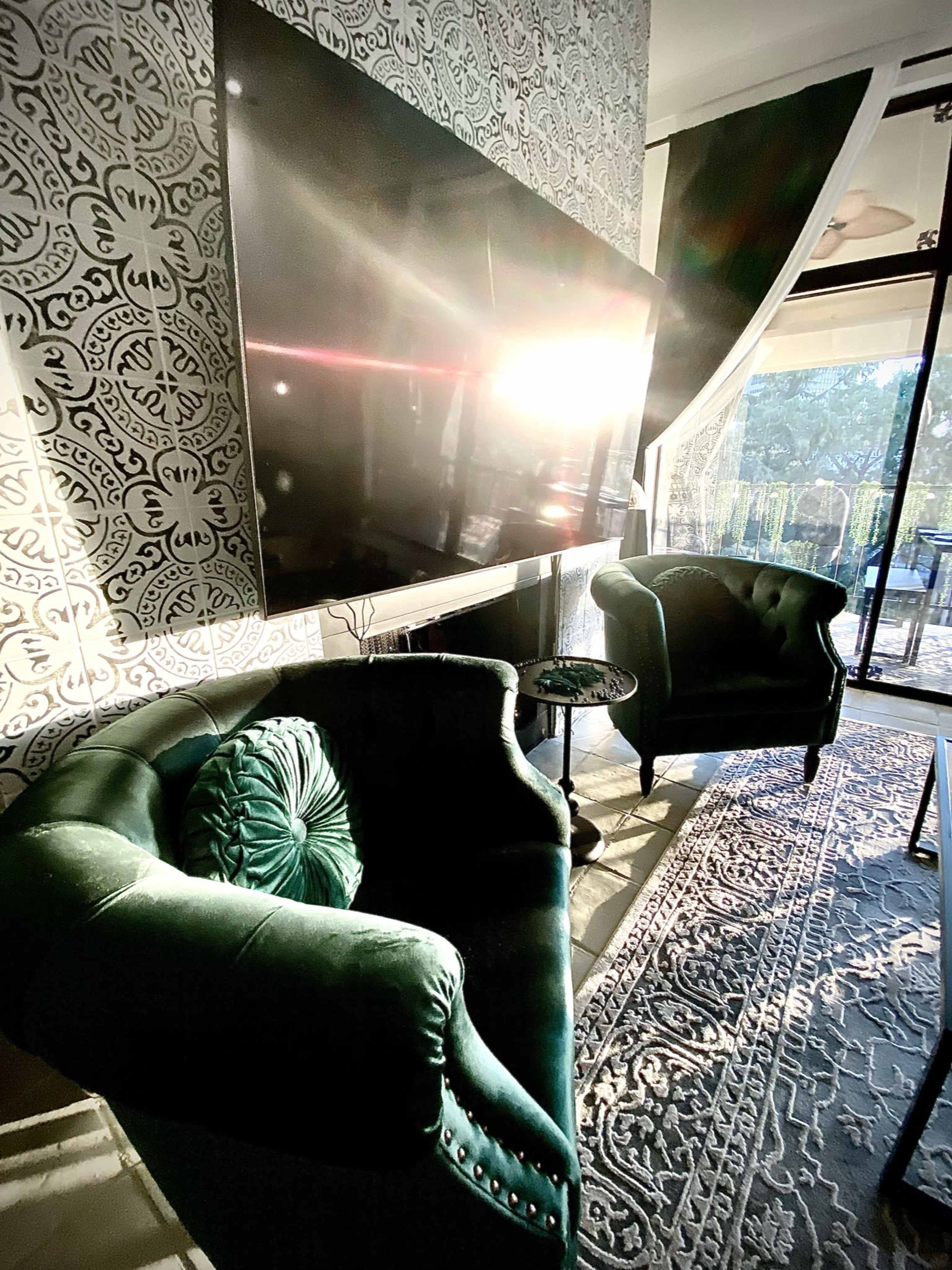 A modern living room with two green velvet chairs, a large flat-screen TV mounted on the wall, and sunlight streaming through a window.