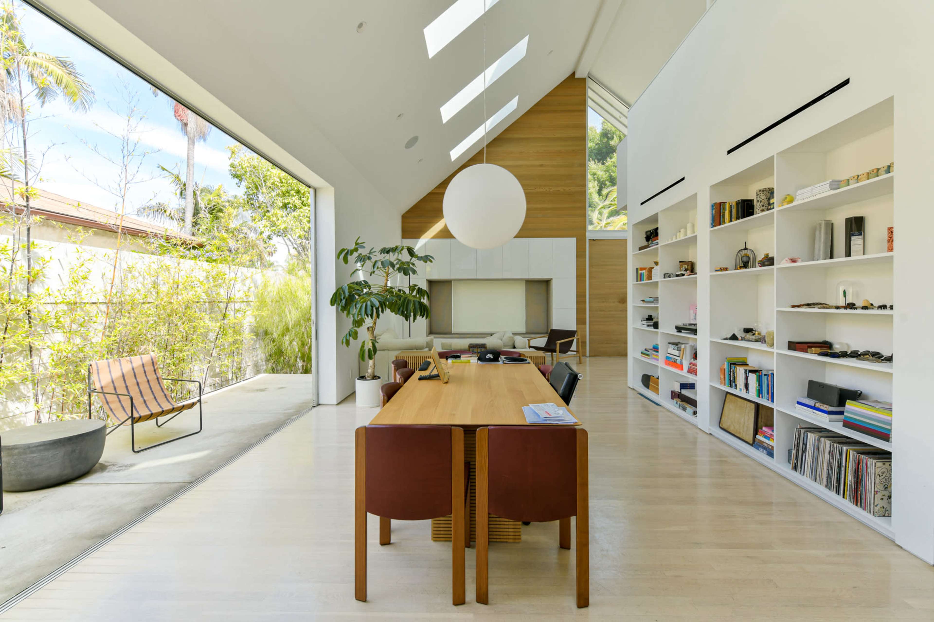 A spacious modern living area features a dining table surrounded by chairs, large windows, and built-in bookshelves.