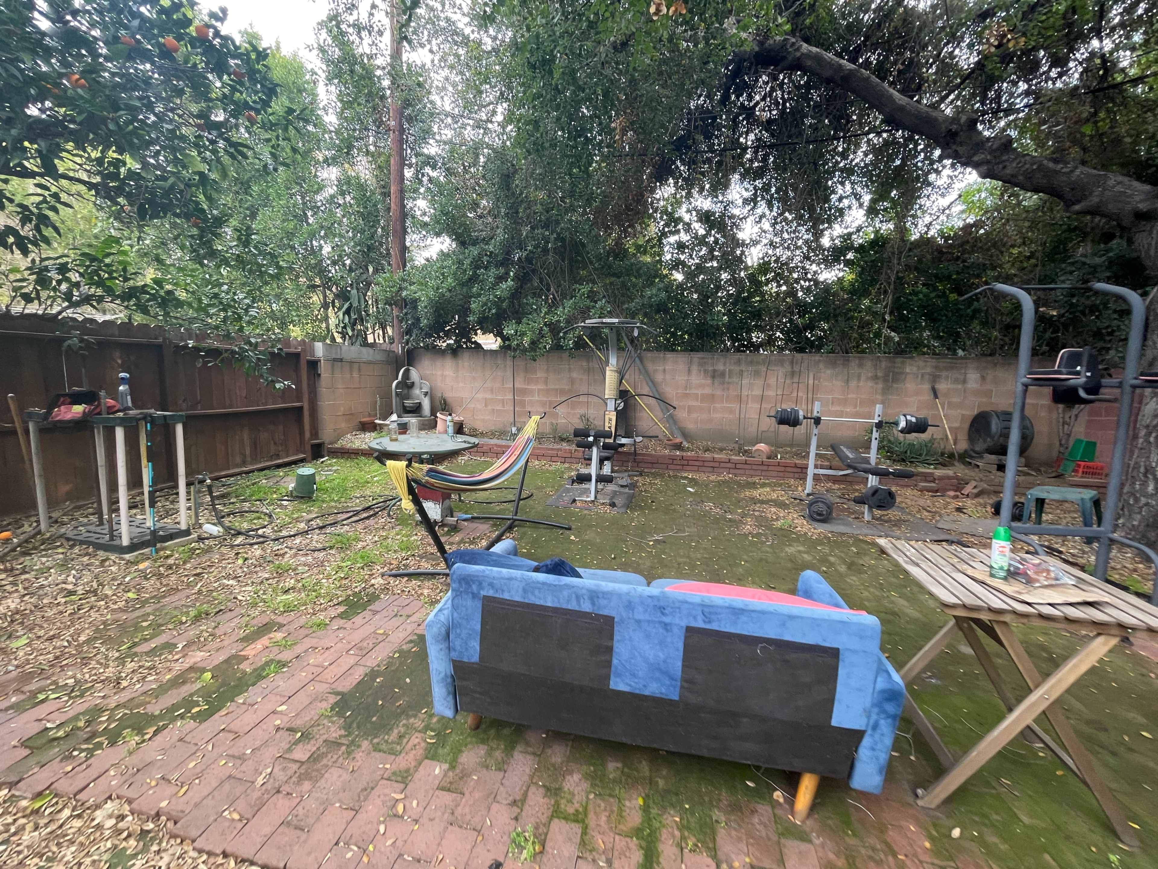 The image shows a backyard with exercise equipment, a hammock, a sofa, and a few garden chairs on a brick patio surrounded by trees.