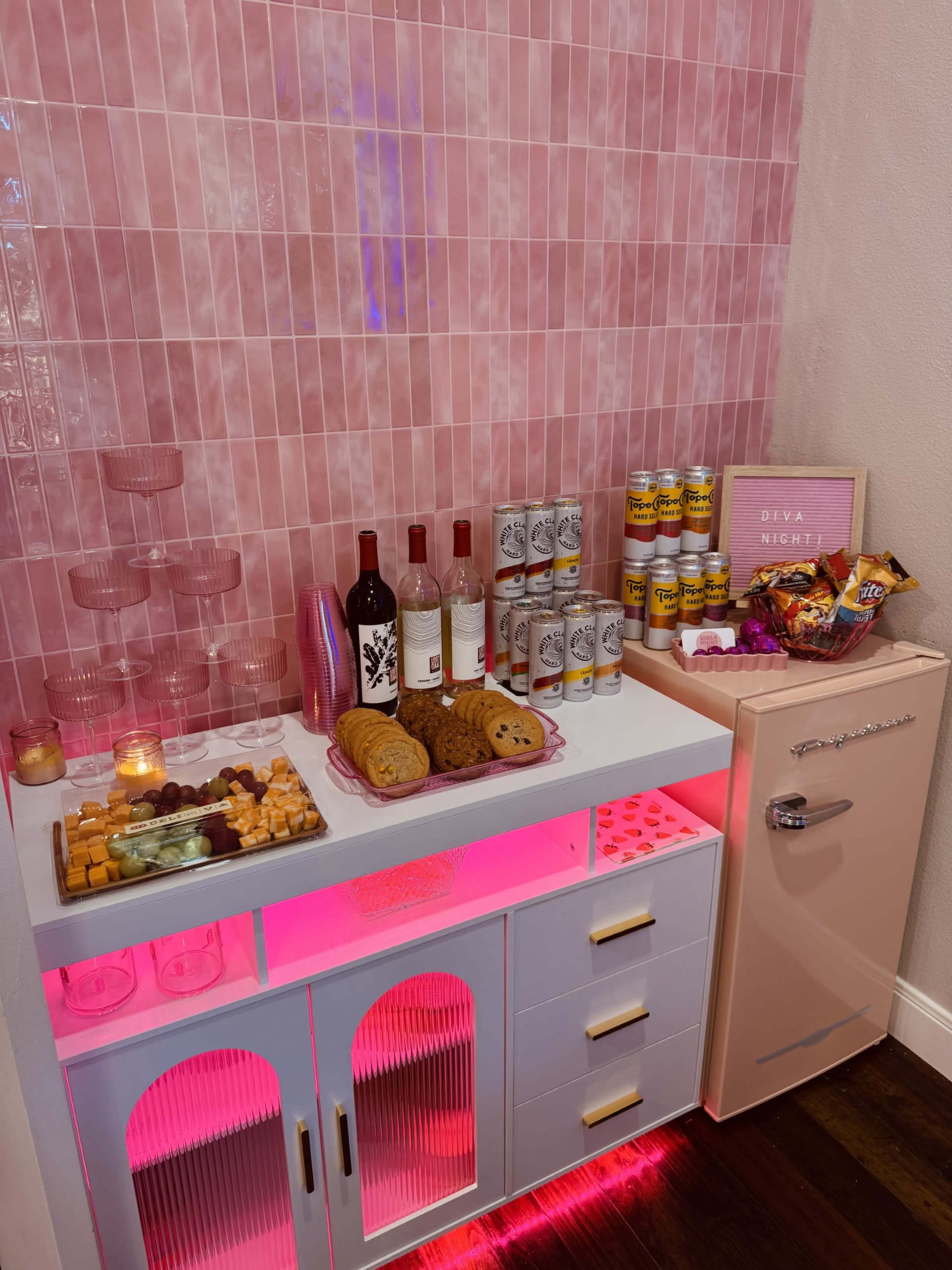 The image shows a stylish beverage and snack station with a pink tiled background, featuring wine bottles, cans, cookies, and a retro-style mini fridge.