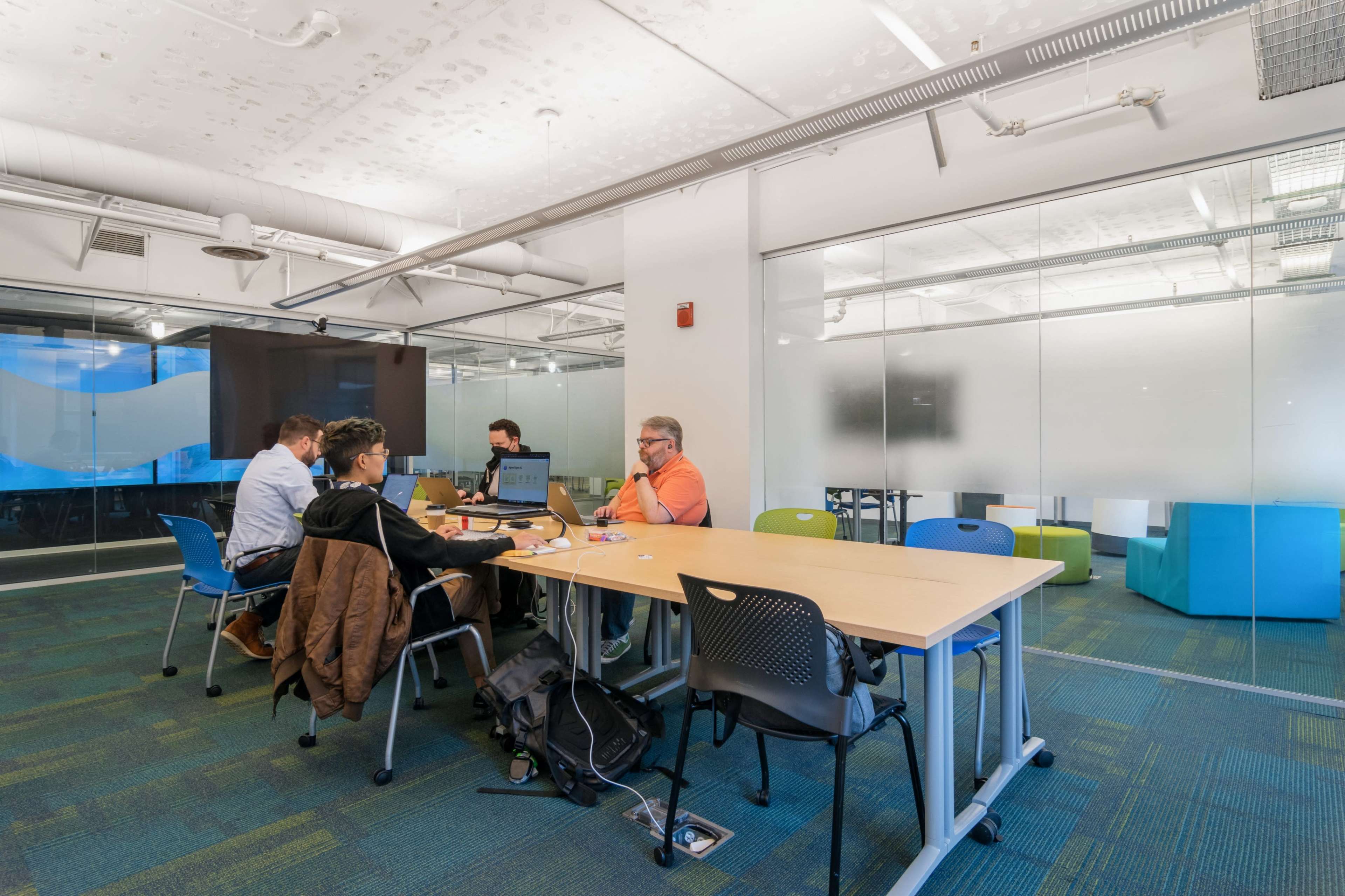 A group of six people collaborates around a large table in a modern office space with large windows and colorful seating areas.