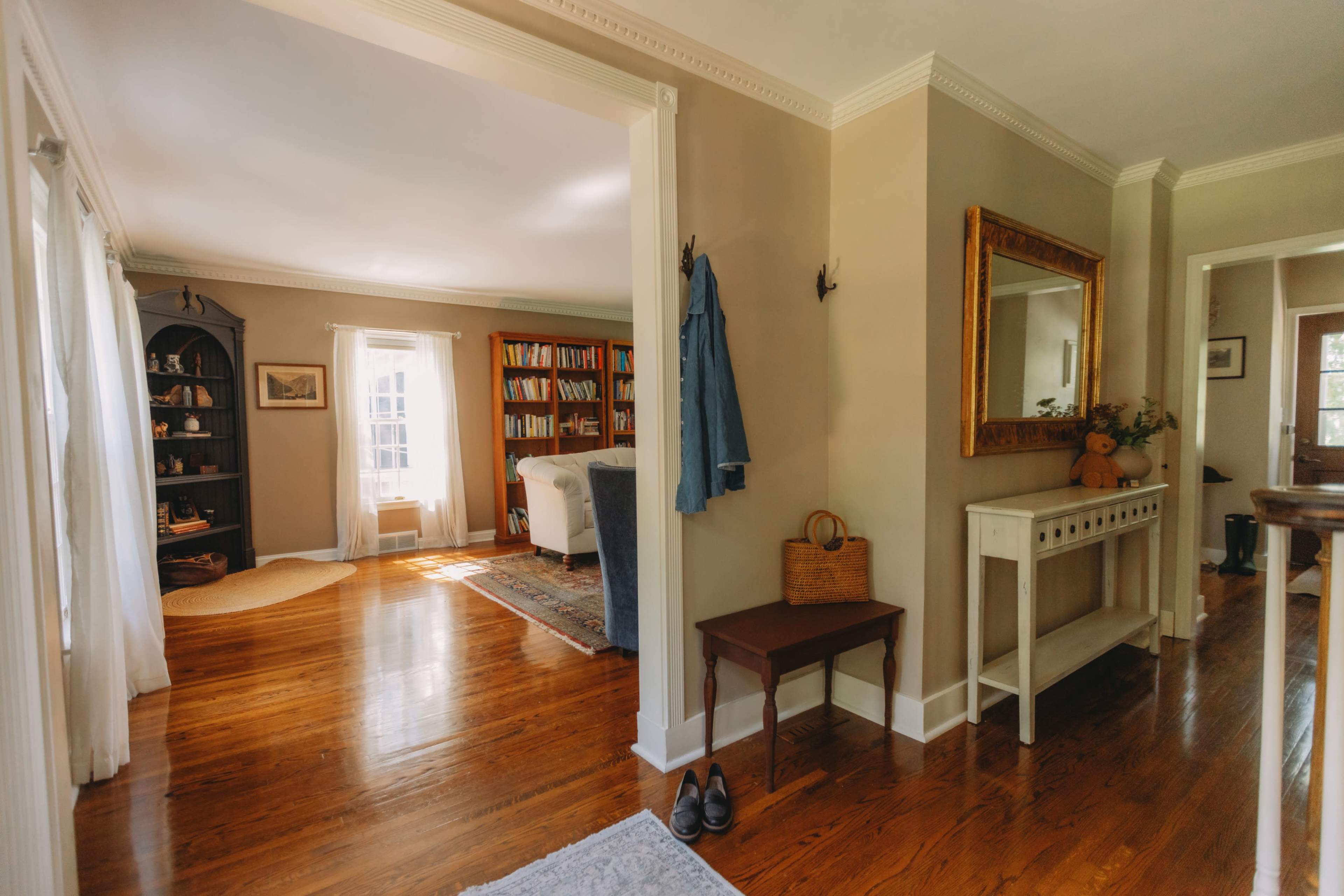 The image shows a sunlit hallway leading to a cozy living area filled with bookshelves and a sofa, framed by white walls and polished wooden floors.