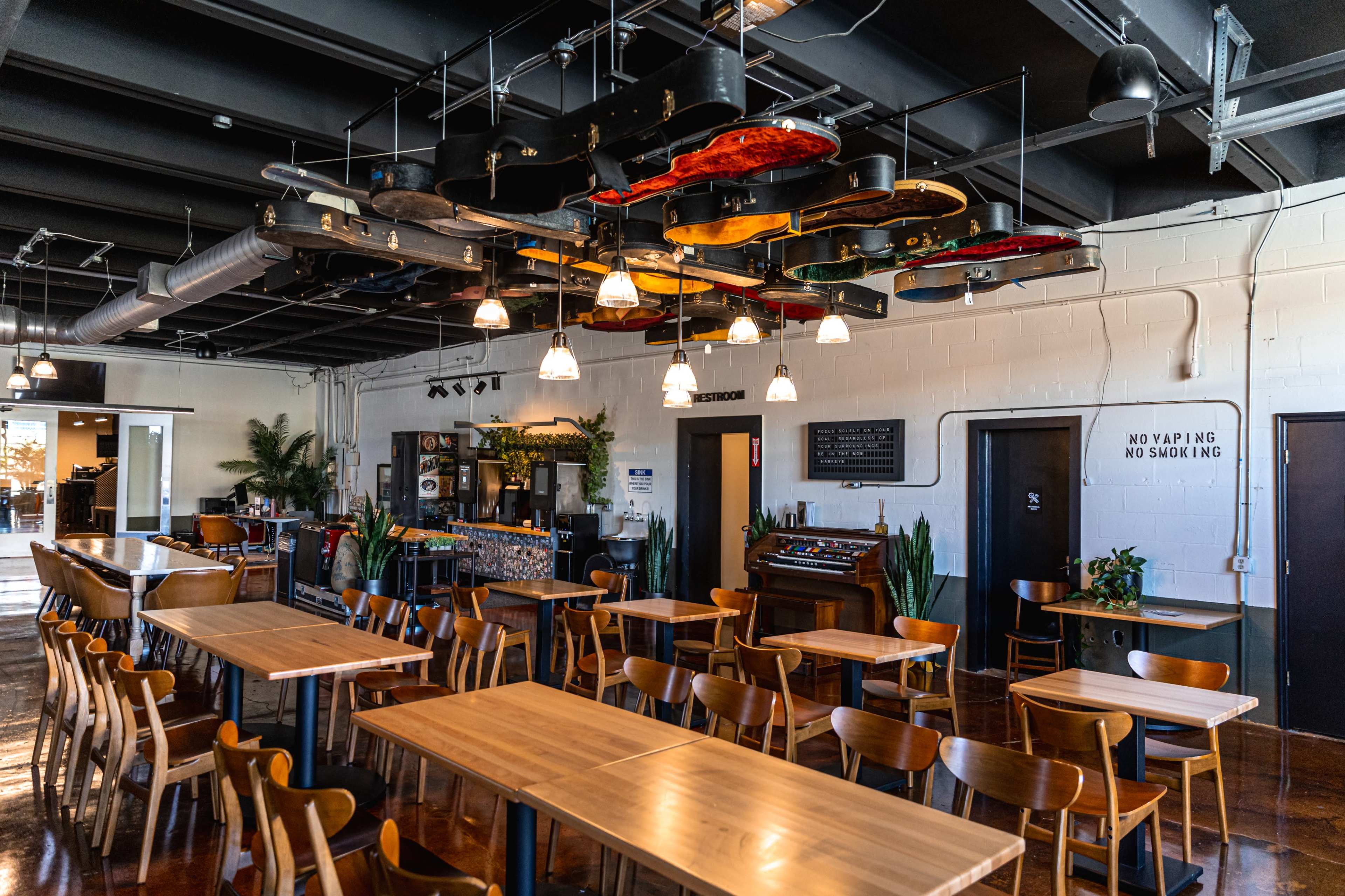 The image shows a spacious restaurant interior with wooden tables and chairs, featuring a ceiling adorned with hanging guitars and several plants throughout the space.