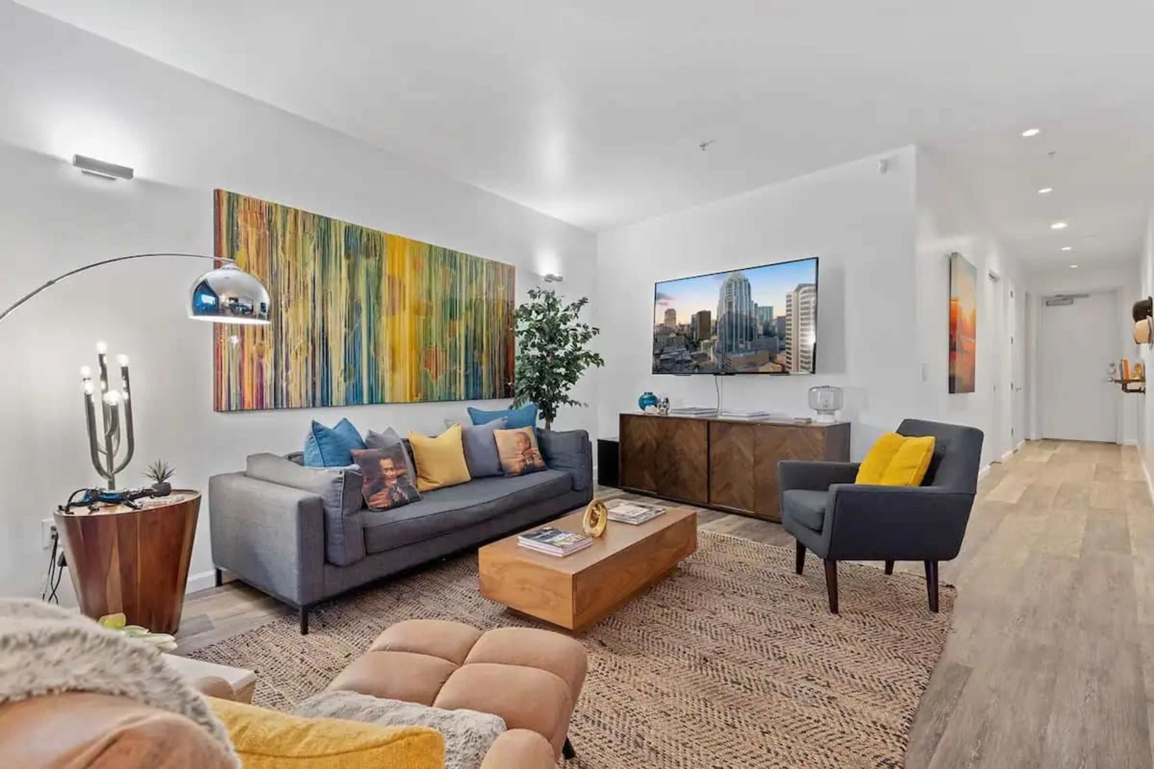 A modern living room featuring a gray sofa with colorful pillows, a wooden coffee table, and a television mounted on a white wall, complemented by vibrant artwork.