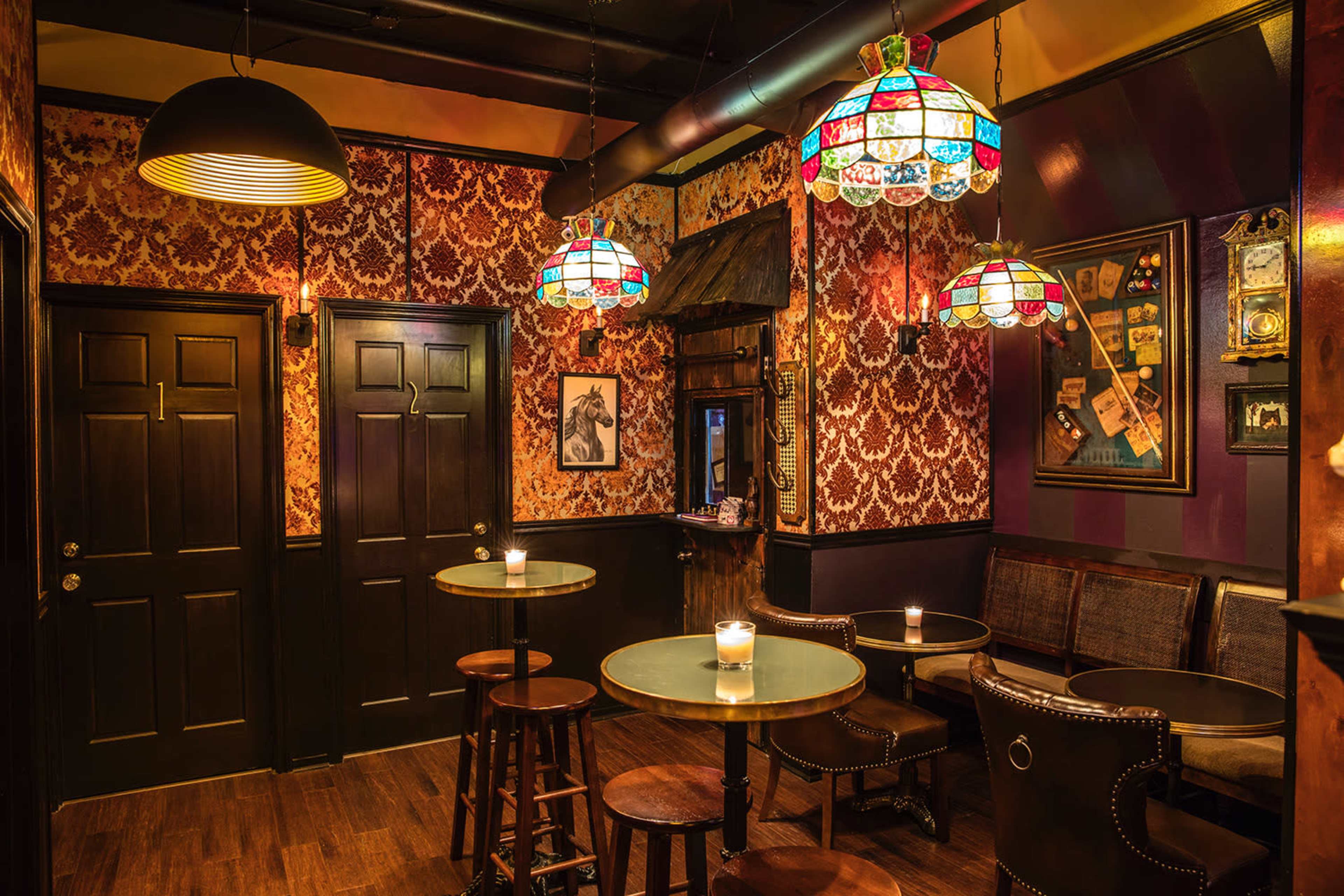 A dimly lit bar with ornate wallpaper, round tables, and colorful stained-glass light fixtures.