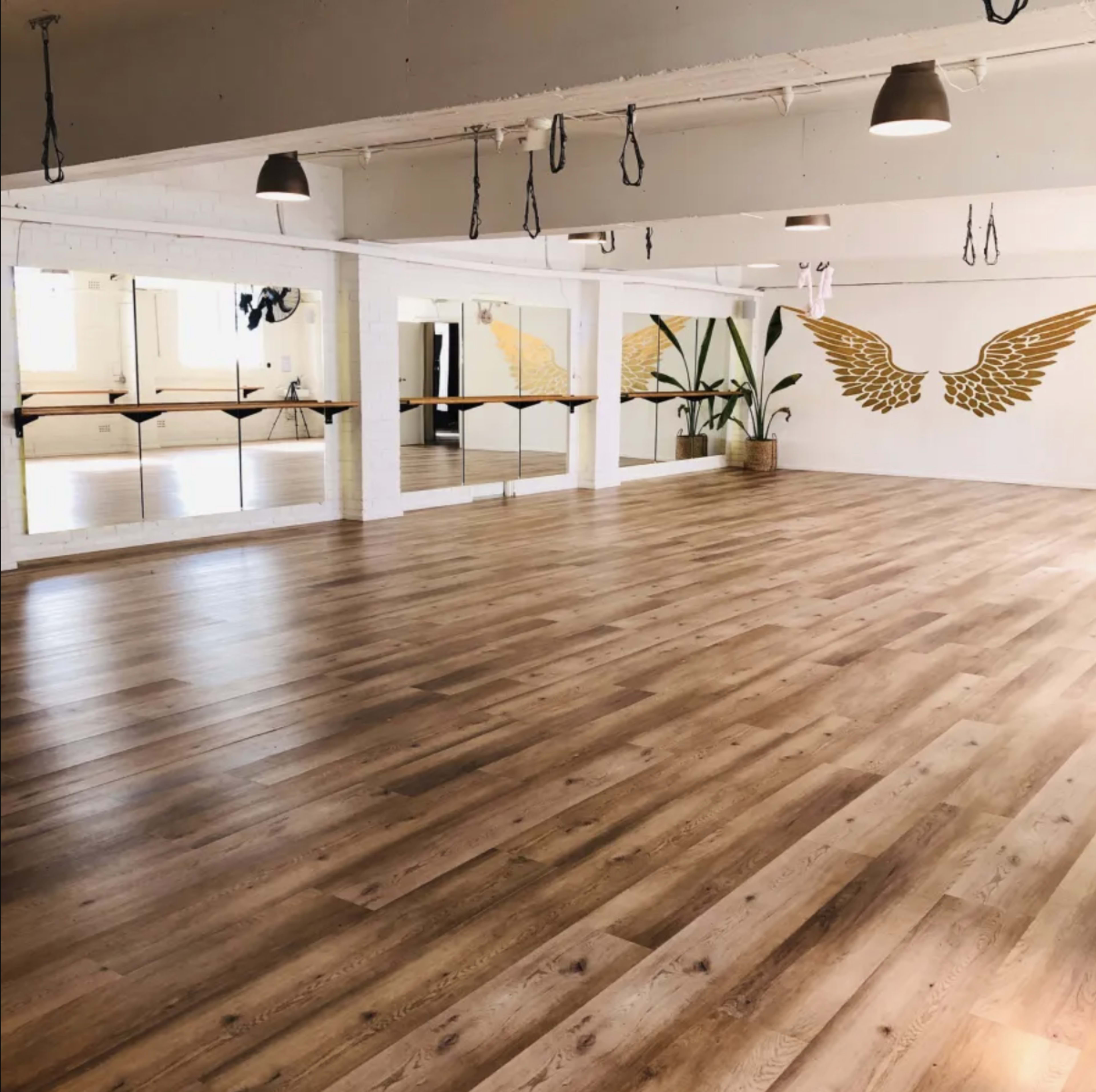 The image shows a spacious fitness studio with wooden flooring, large mirrors, and a wall decorated with golden wings.