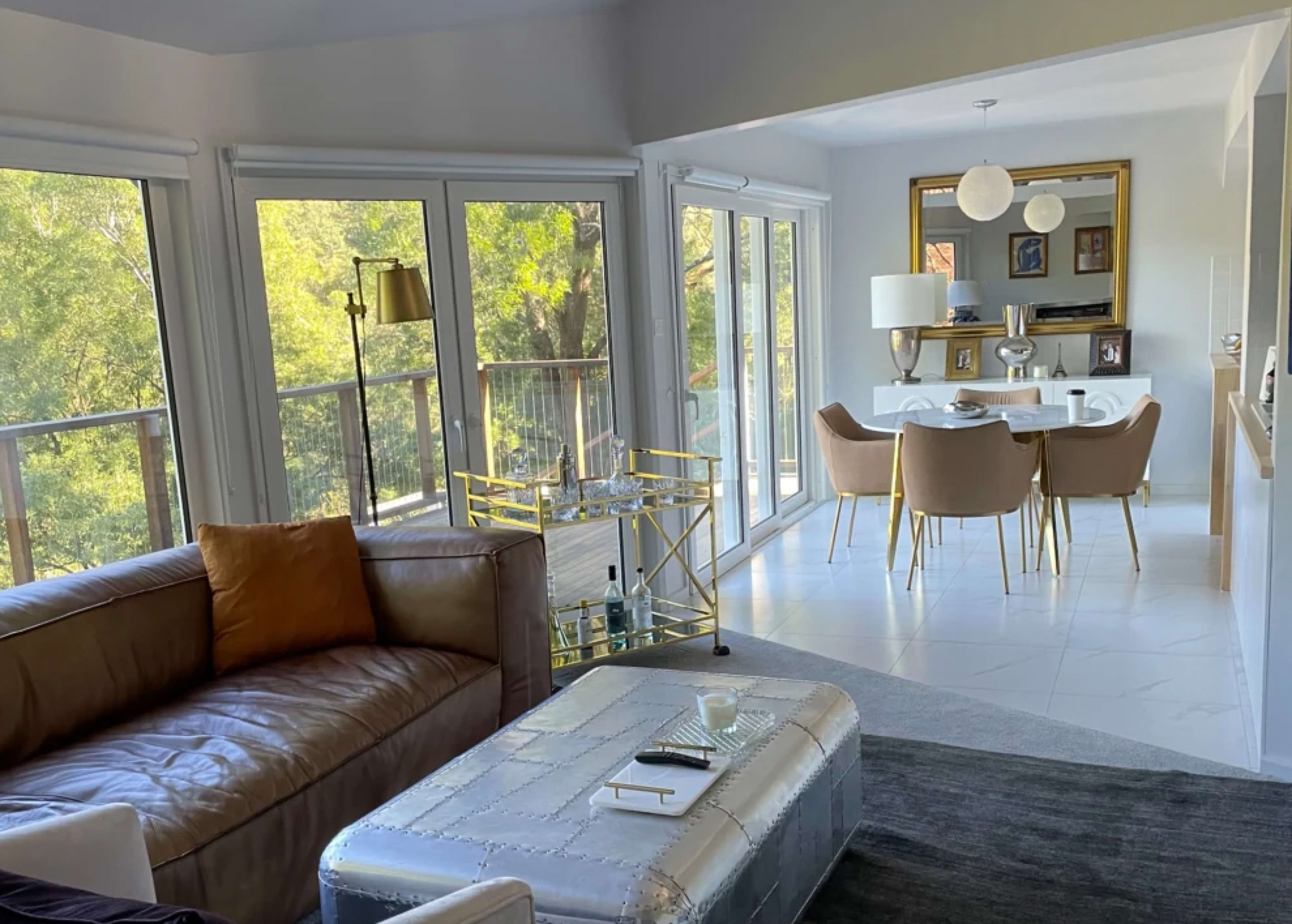 A spacious living area features a brown leather sofa with an orange pillow, a glass coffee table, and a dining space with a white table and beige chairs, all set against large windows overlooking greenery.