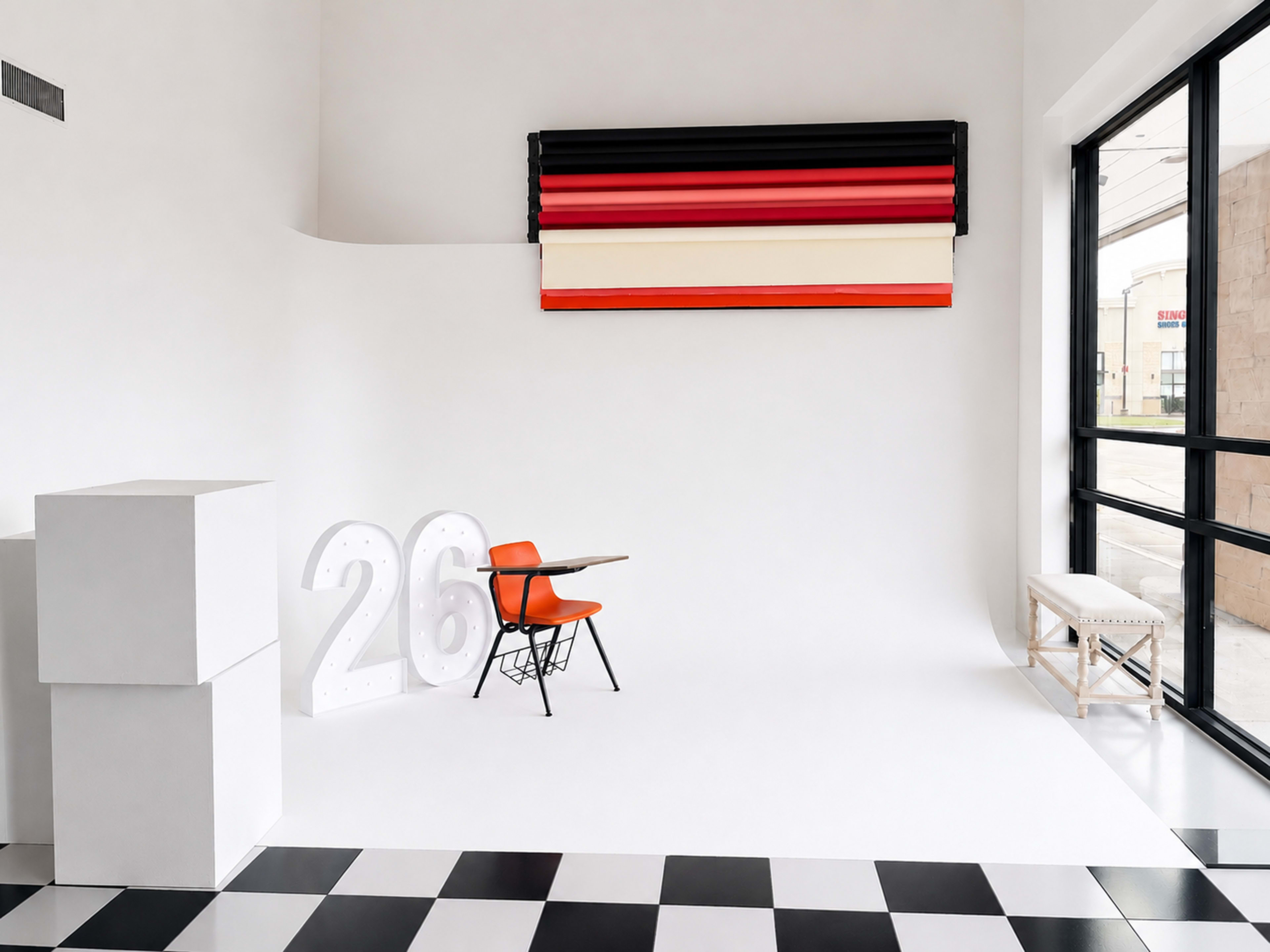 The image shows a modern interior space featuring a black and white checkered floor, a white wall with a colorful roller blind, an orange chair, a small table, and a large illuminated number "26."