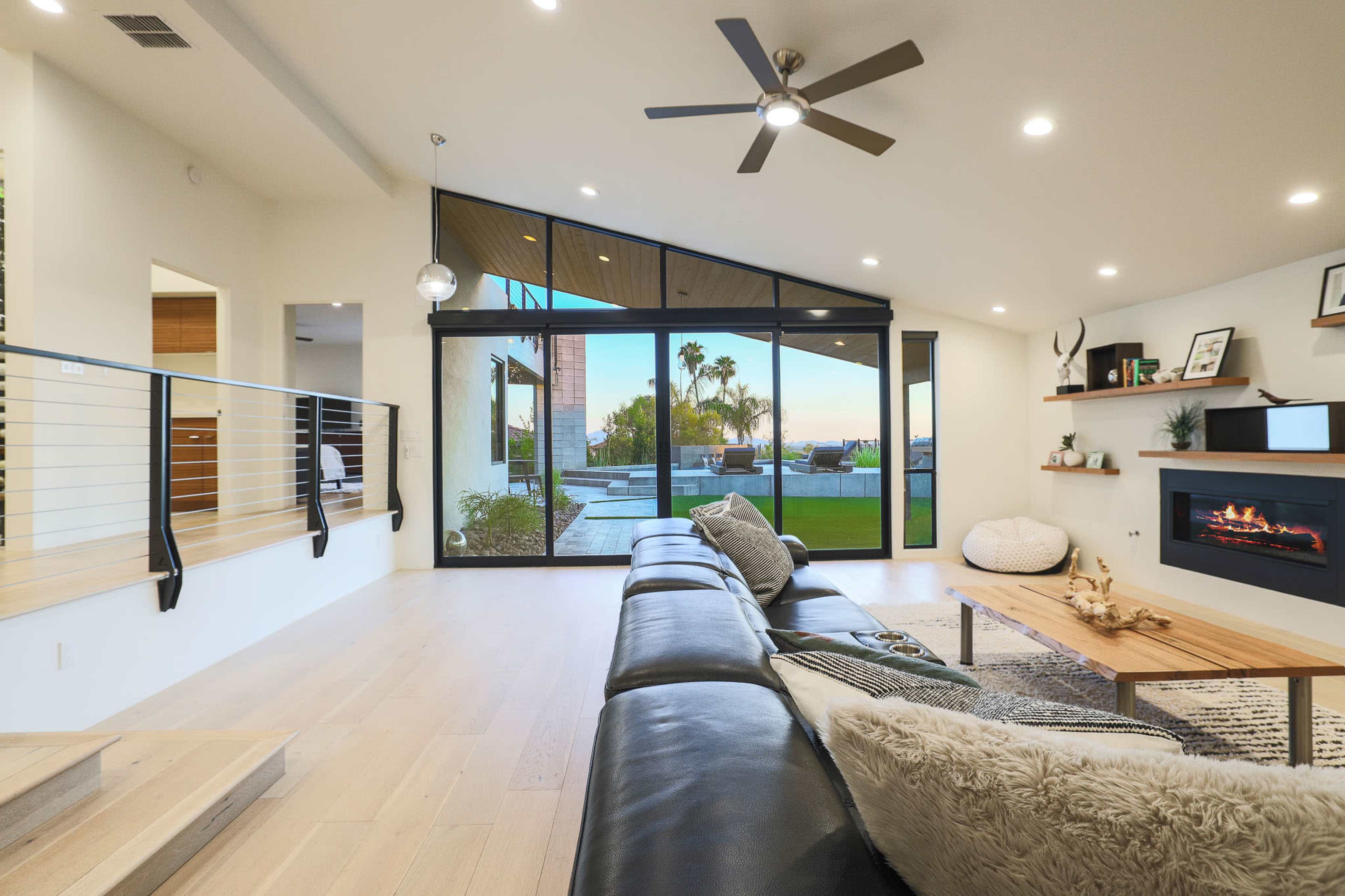 A modern living room with a black sofa, a wooden coffee table, and large glass doors leading to an outdoor area with a view of palm trees.