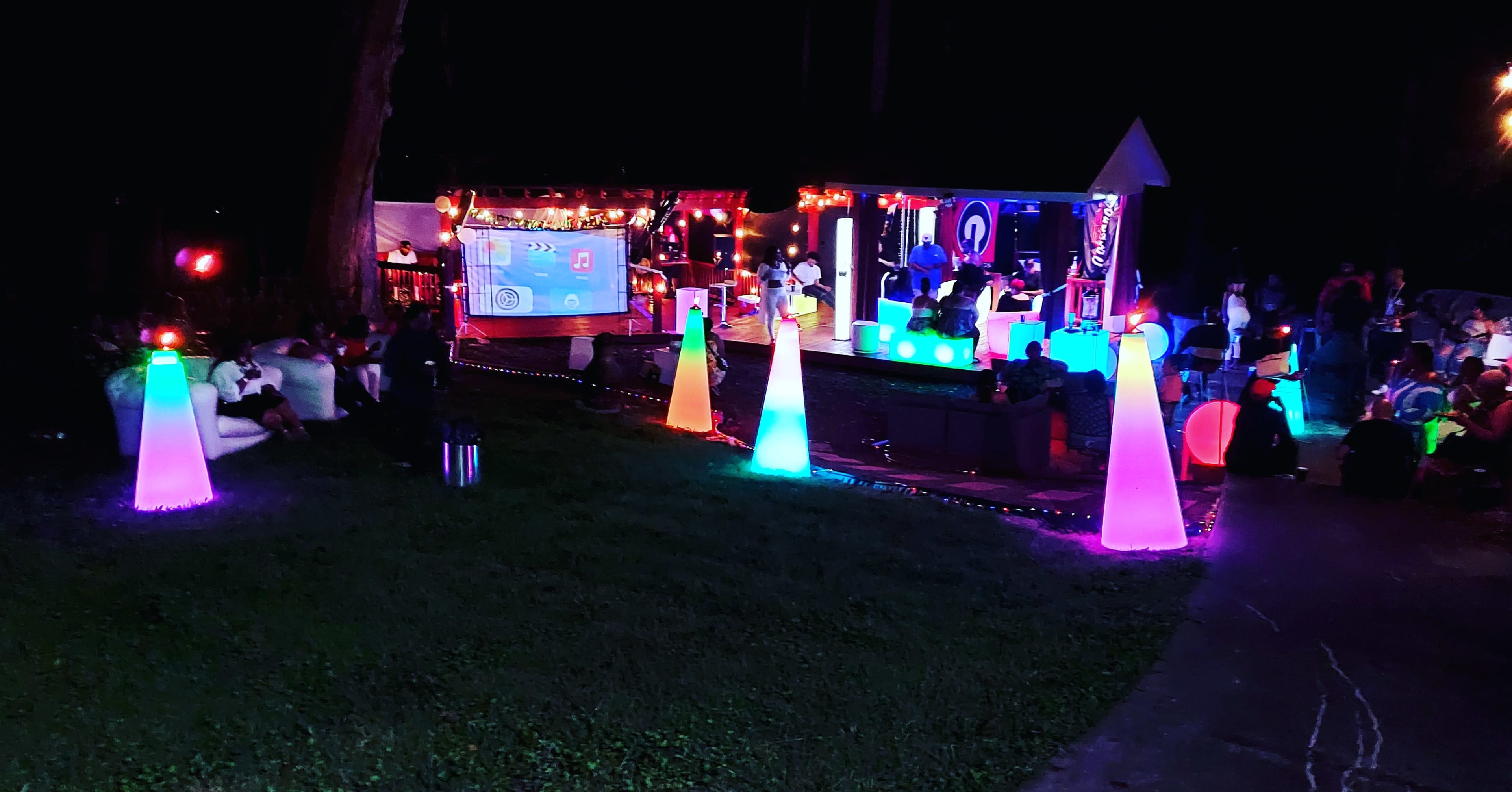 A brightly lit outdoor gathering features colorful illuminated decorations and a stage with a projector, surrounded by guests seated on the grass.