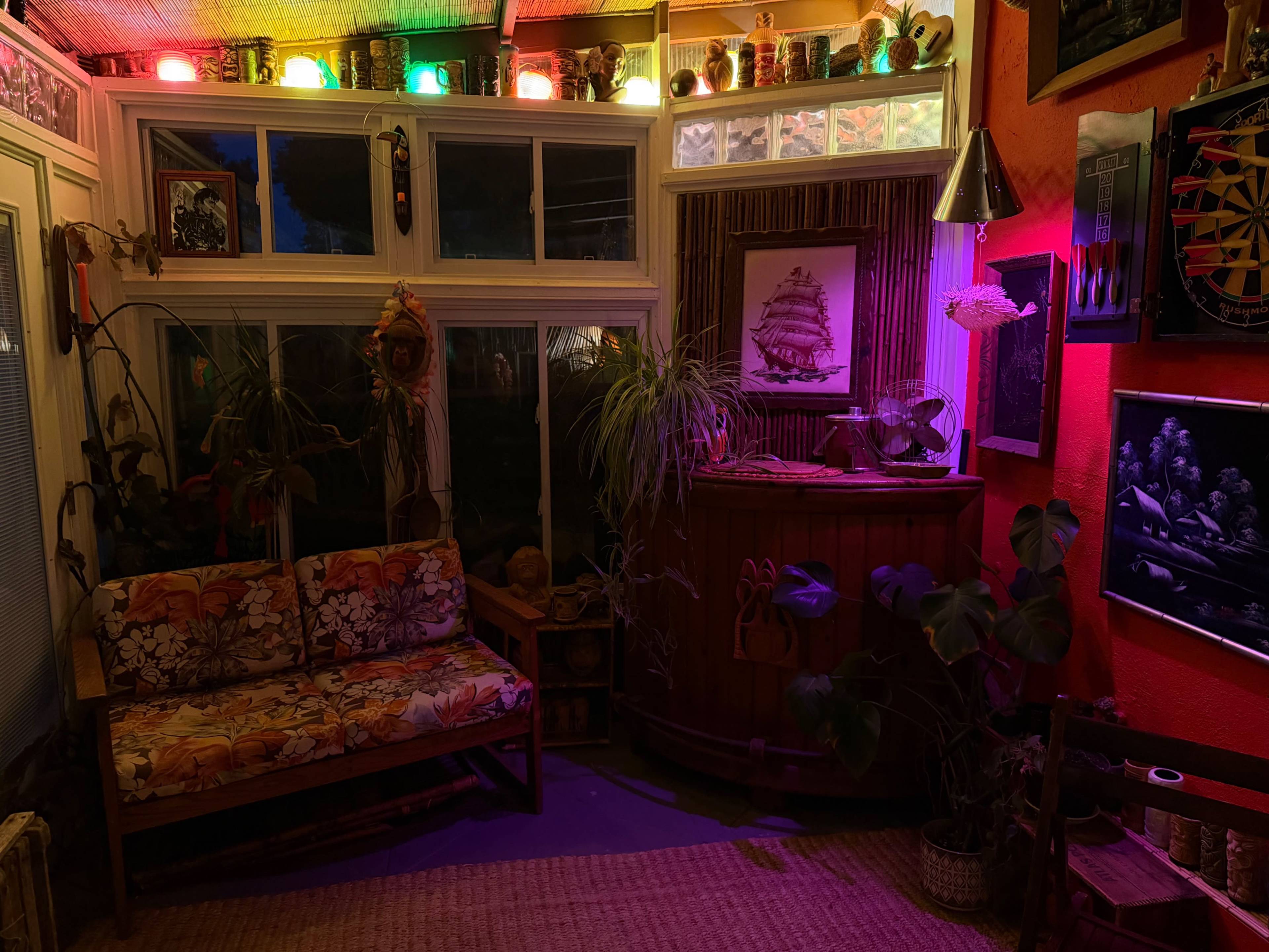 The image shows a cozy indoor space with colorful lights, a floral-patterned sofa, and various plants, illuminated by soft, ambient lighting.