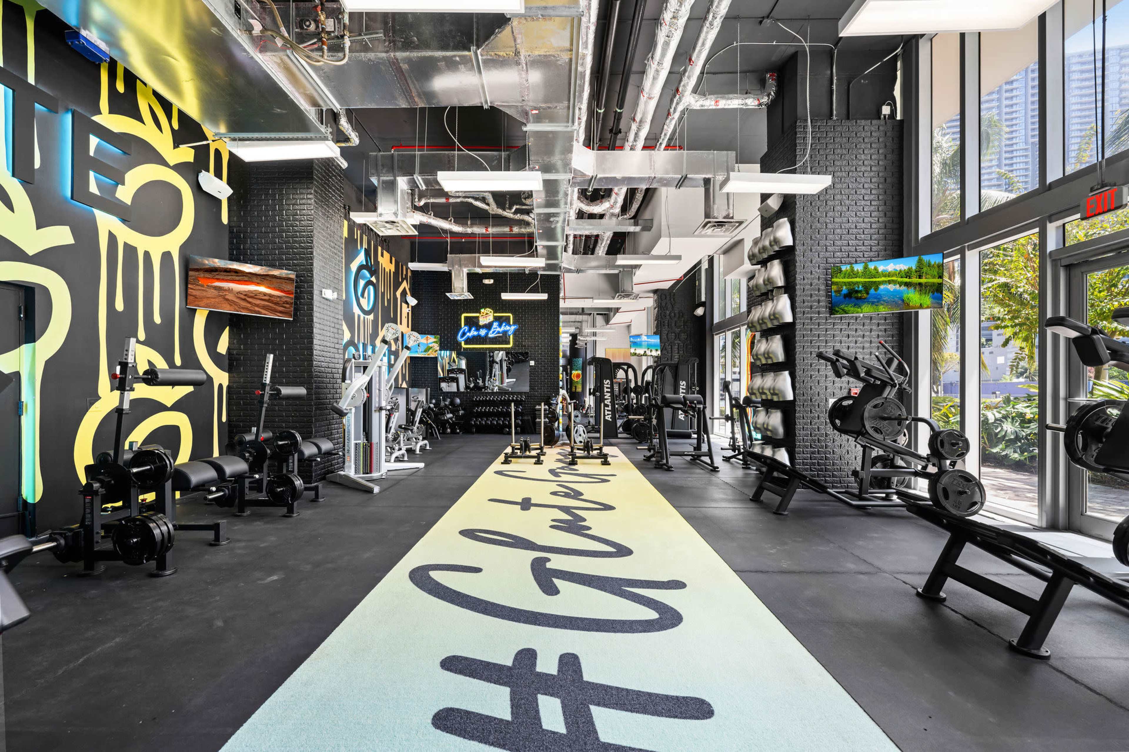 A modern gym interior featuring various workout equipment, neon wall art, and a hashtag design on the floor.
