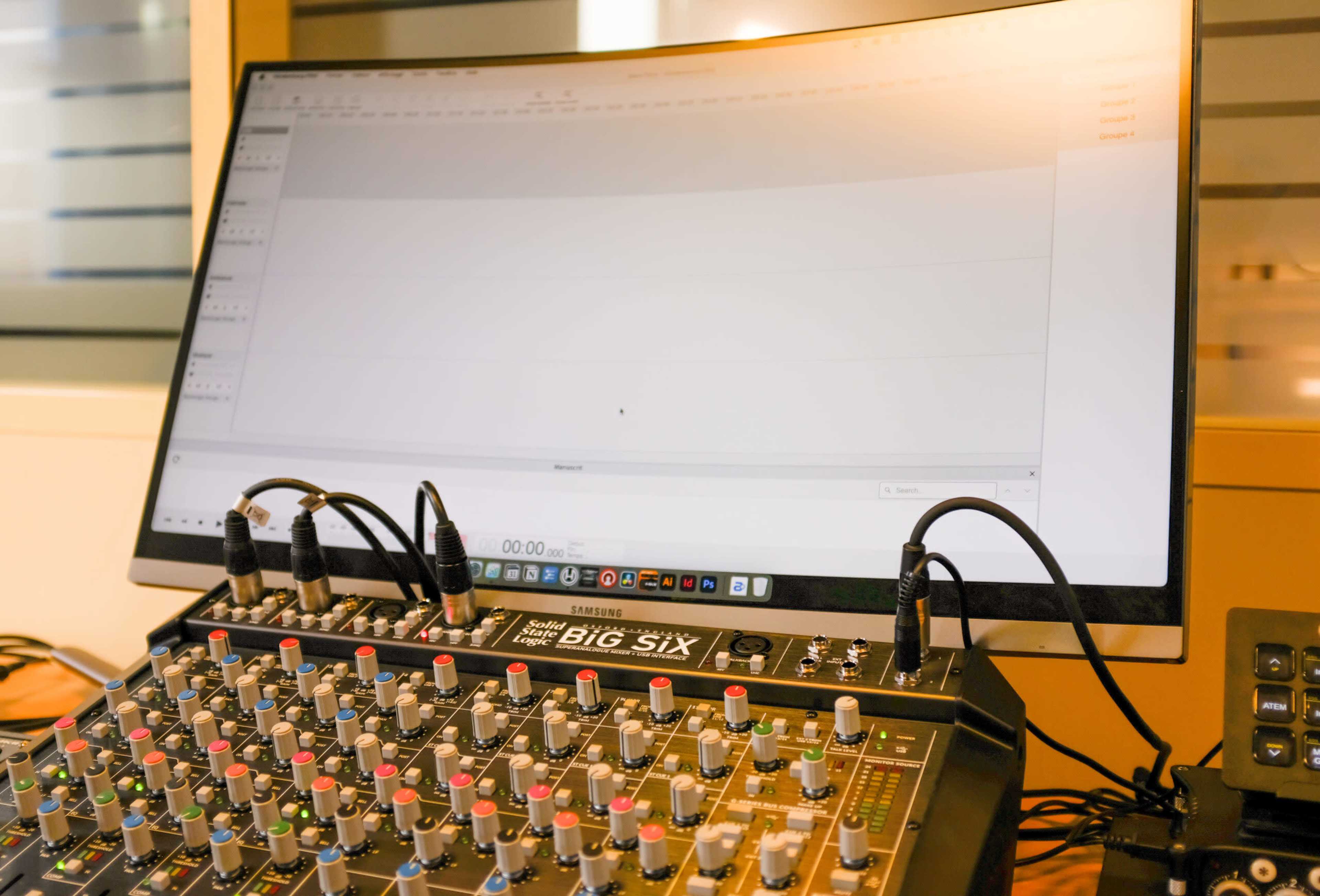 A large monitor displays audio editing software above a mixing console with various knobs and sliders.