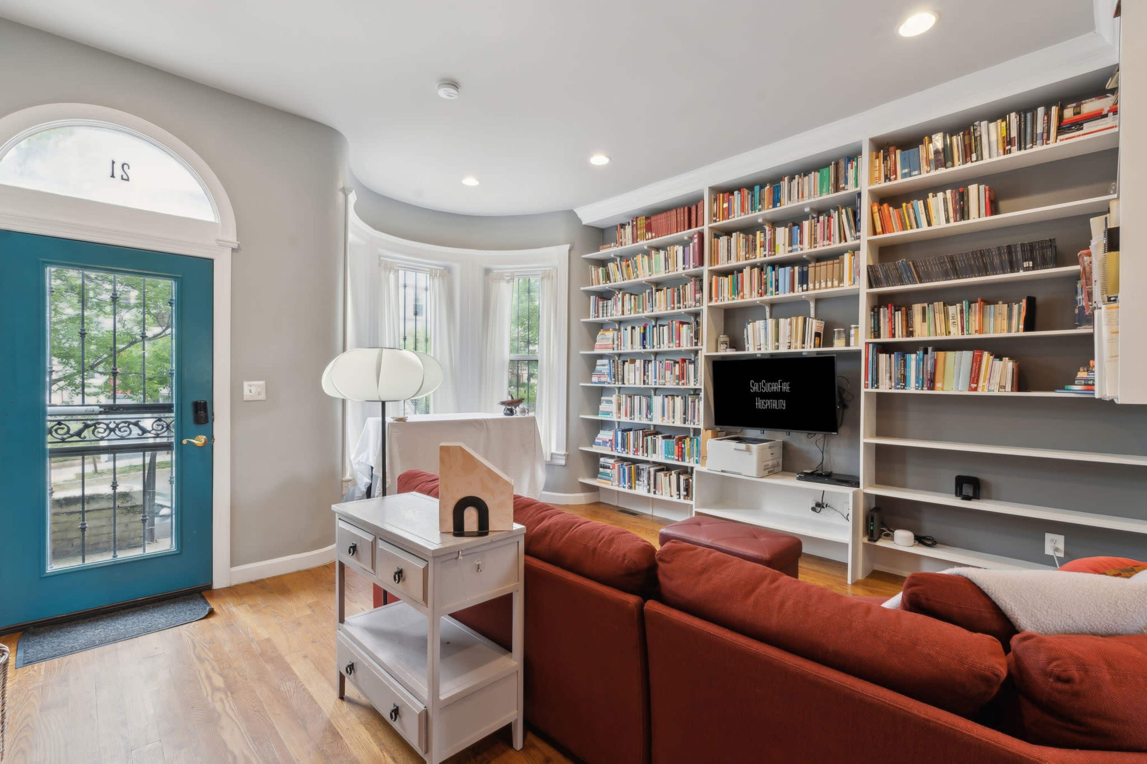The image shows a cozy living room featuring a large bookshelf filled with books, a red sofa, a television, and a blue front door.