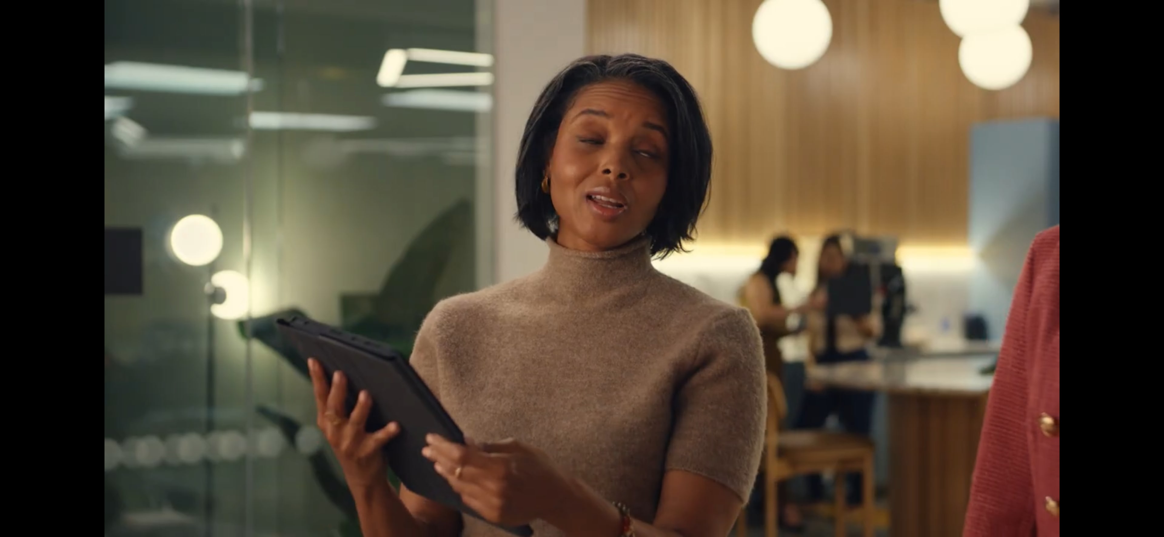 A woman in a beige sweater holds a tablet while speaking in a modern office setting.