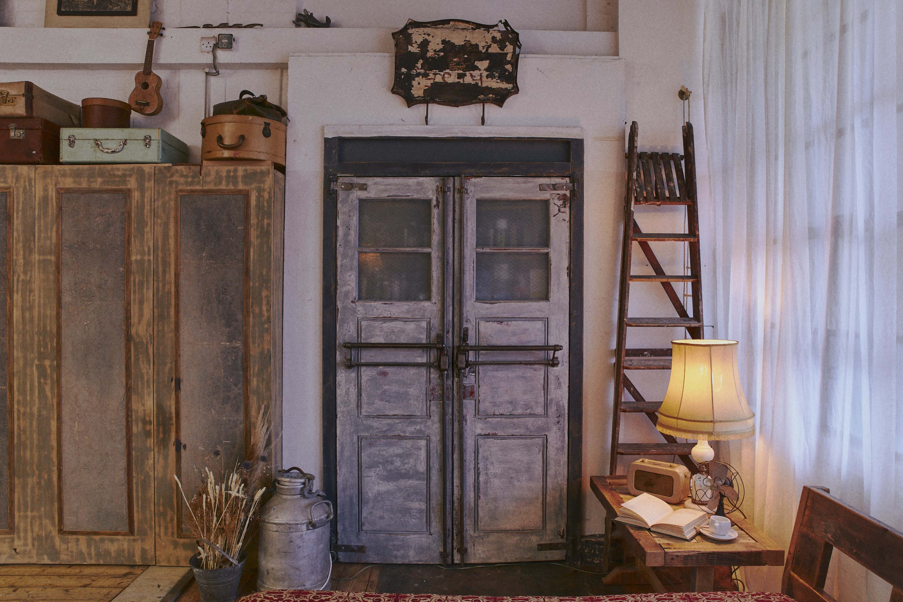 A vintage-style room with a pair of aged double doors, a wooden ladder, a lamp, and various items such as a sewing machine and storage boxes arranged around the space.
