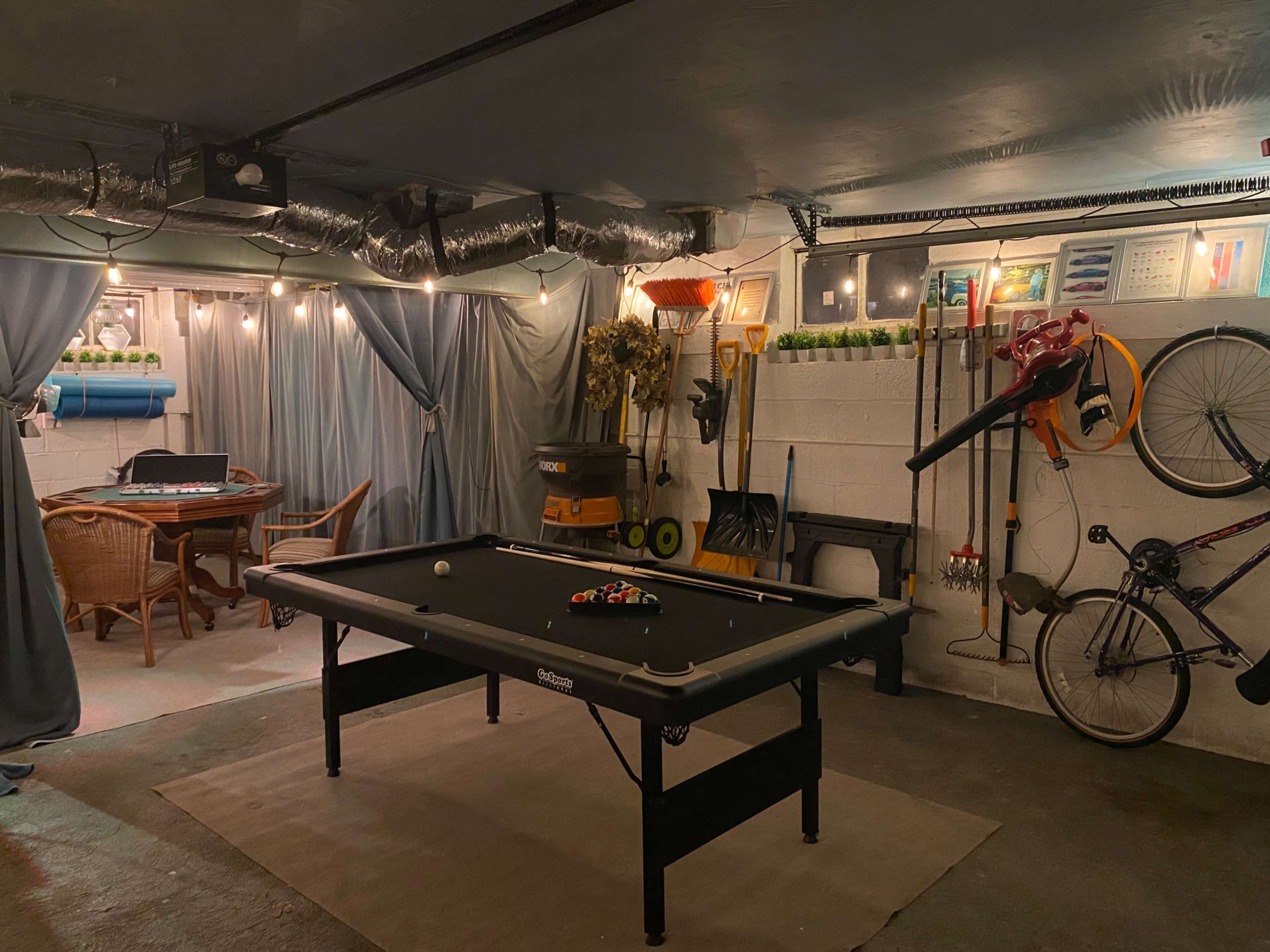 The image shows a dimly lit basement recreation area with a pool table, a small dining table, and various tools and bicycles stored on the walls.