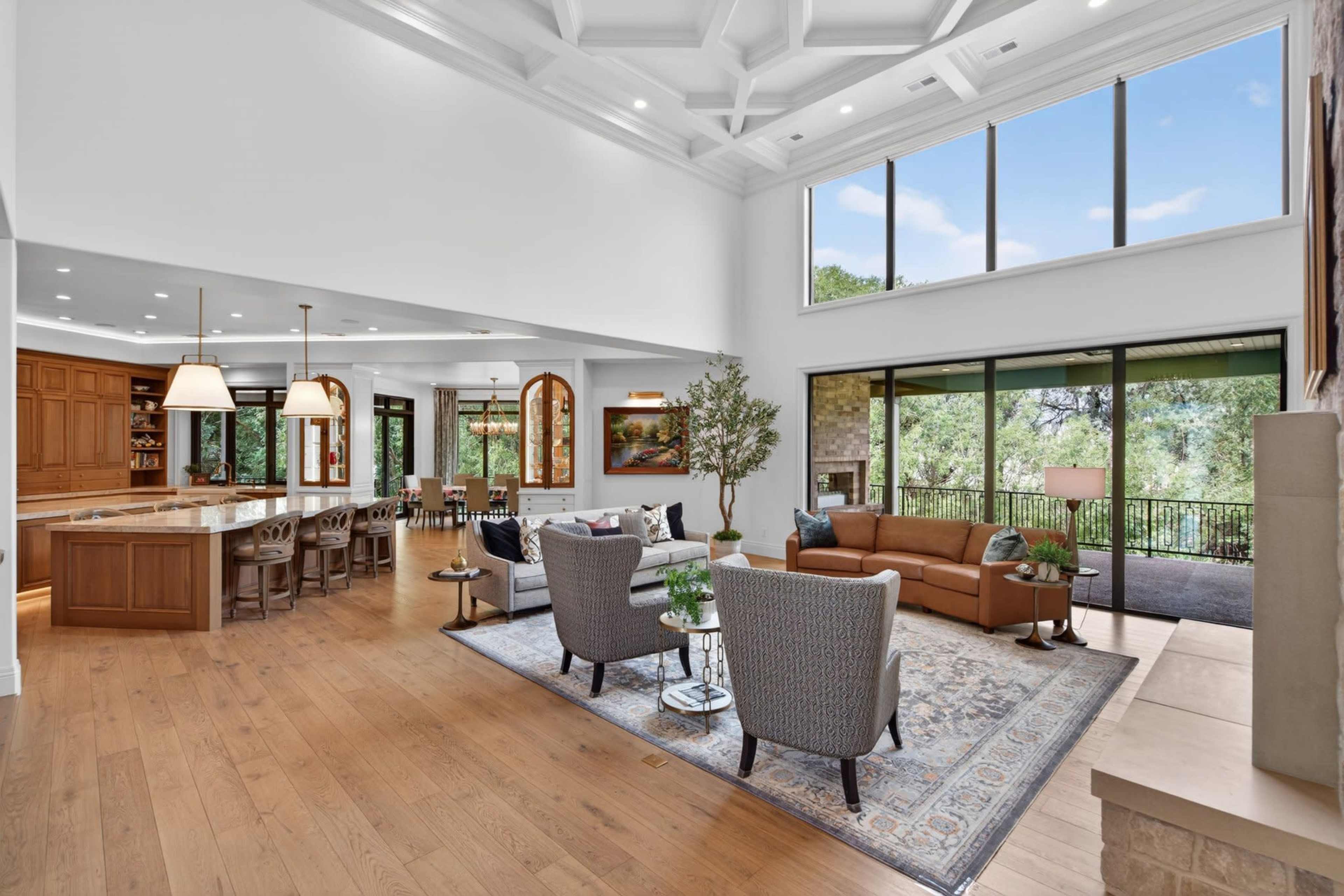 The image displays a spacious living area featuring a high ceiling, large windows, and a blend of seating areas that overlook a natural landscape.