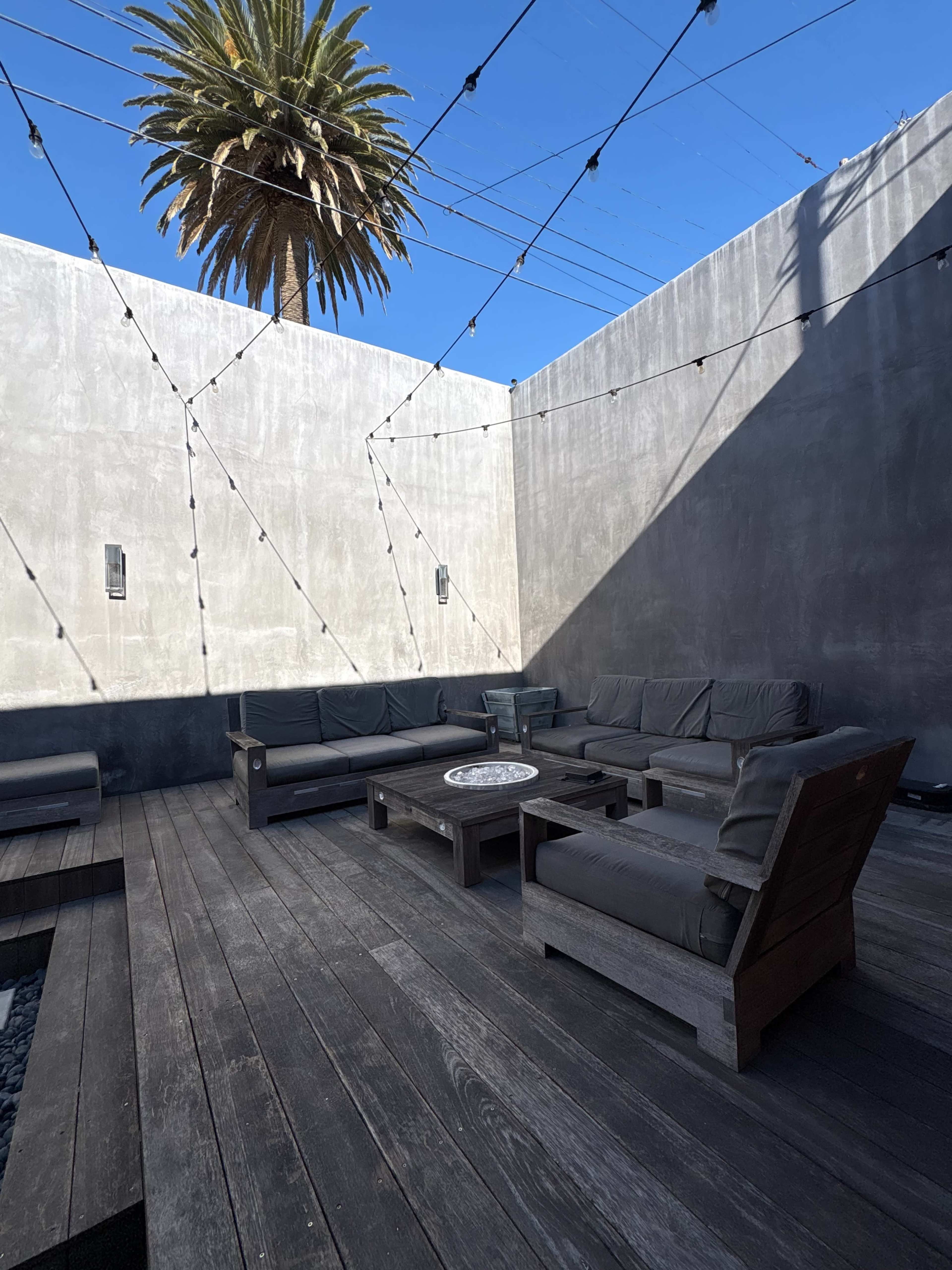 The image features a modern outdoor lounge area with dark wooden decking, comfortable gray seating, and a tall palm tree in the background under a clear blue sky.