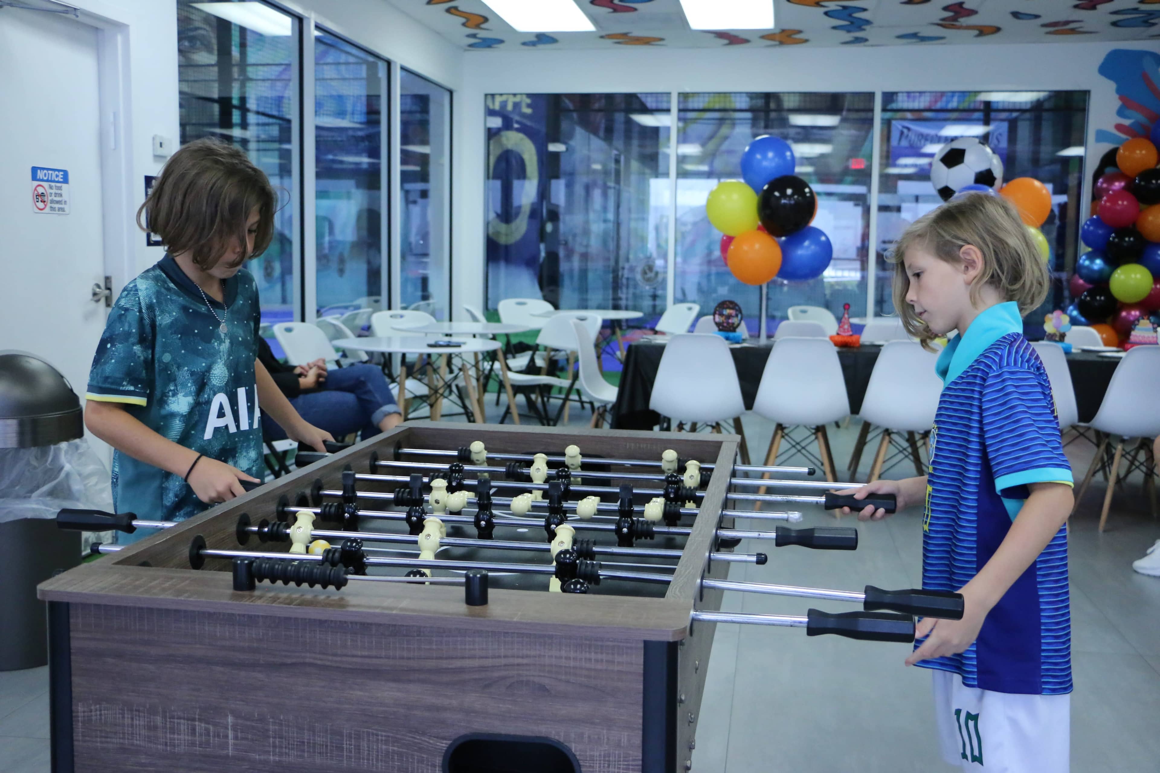 Two children play foosball in a brightly decorated recreational area with colorful balloons in the background.