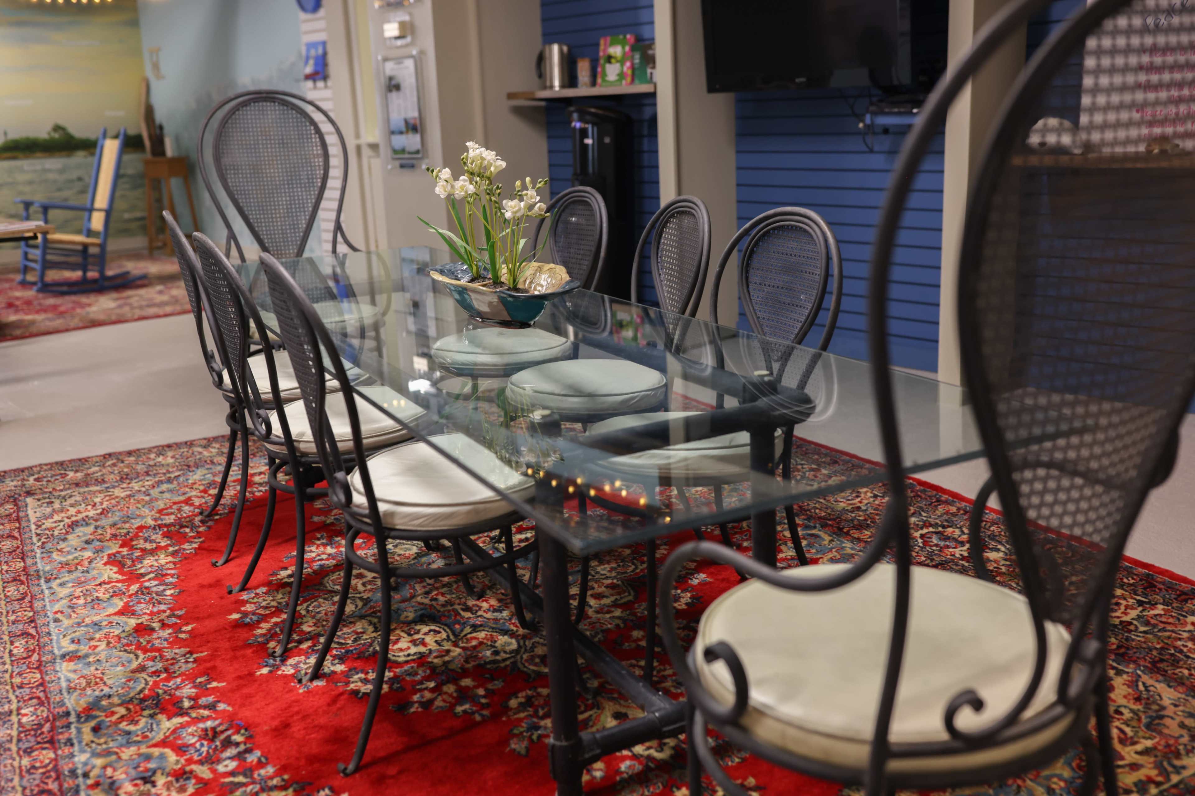 A glass dining table surrounded by eight chairs sits on a decorative red rug in a well-lit room.