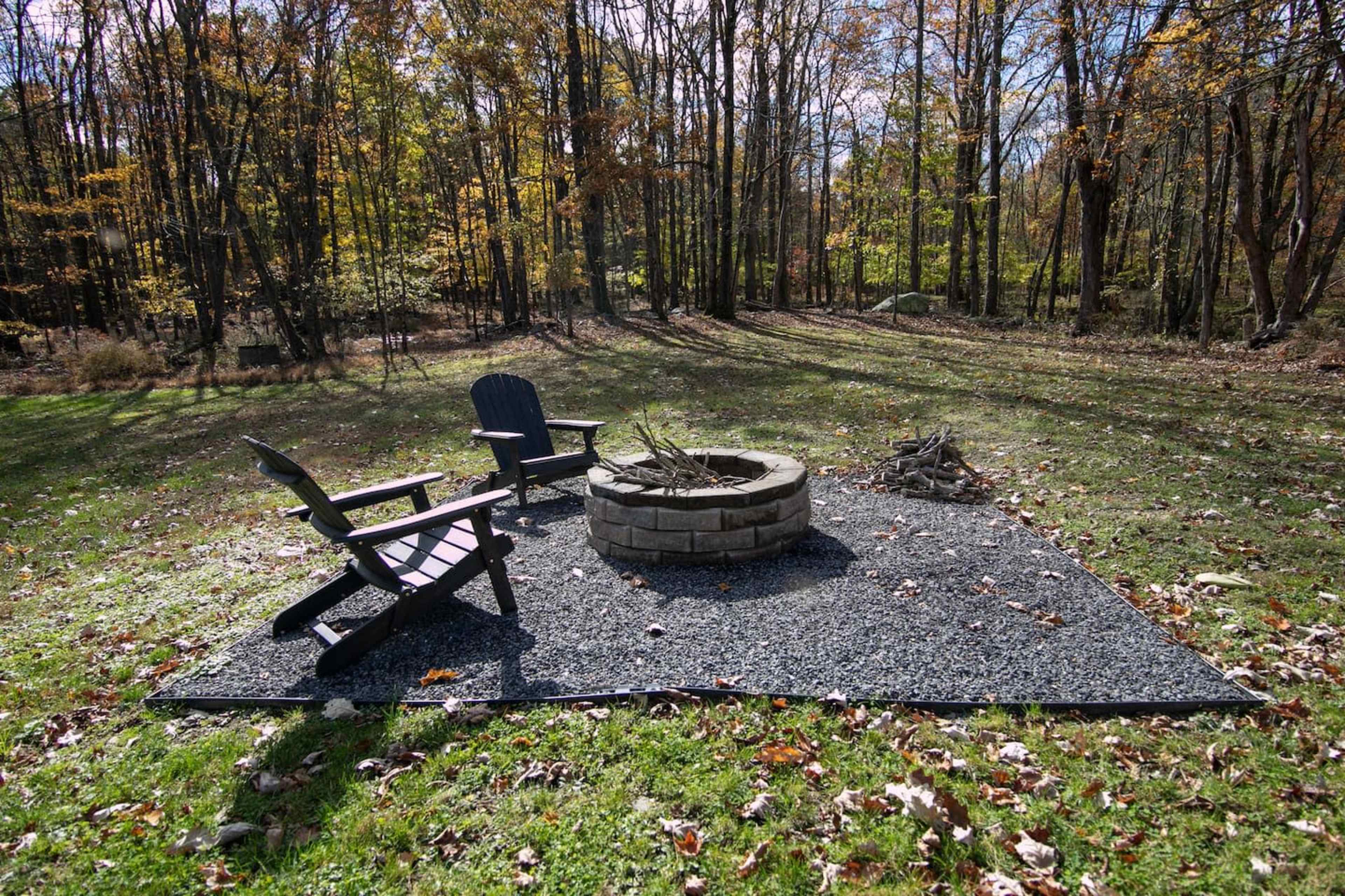 A circular stone fire pit is surrounded by two wooden chairs on a gravel area in a wooded outdoor setting with autumn foliage.