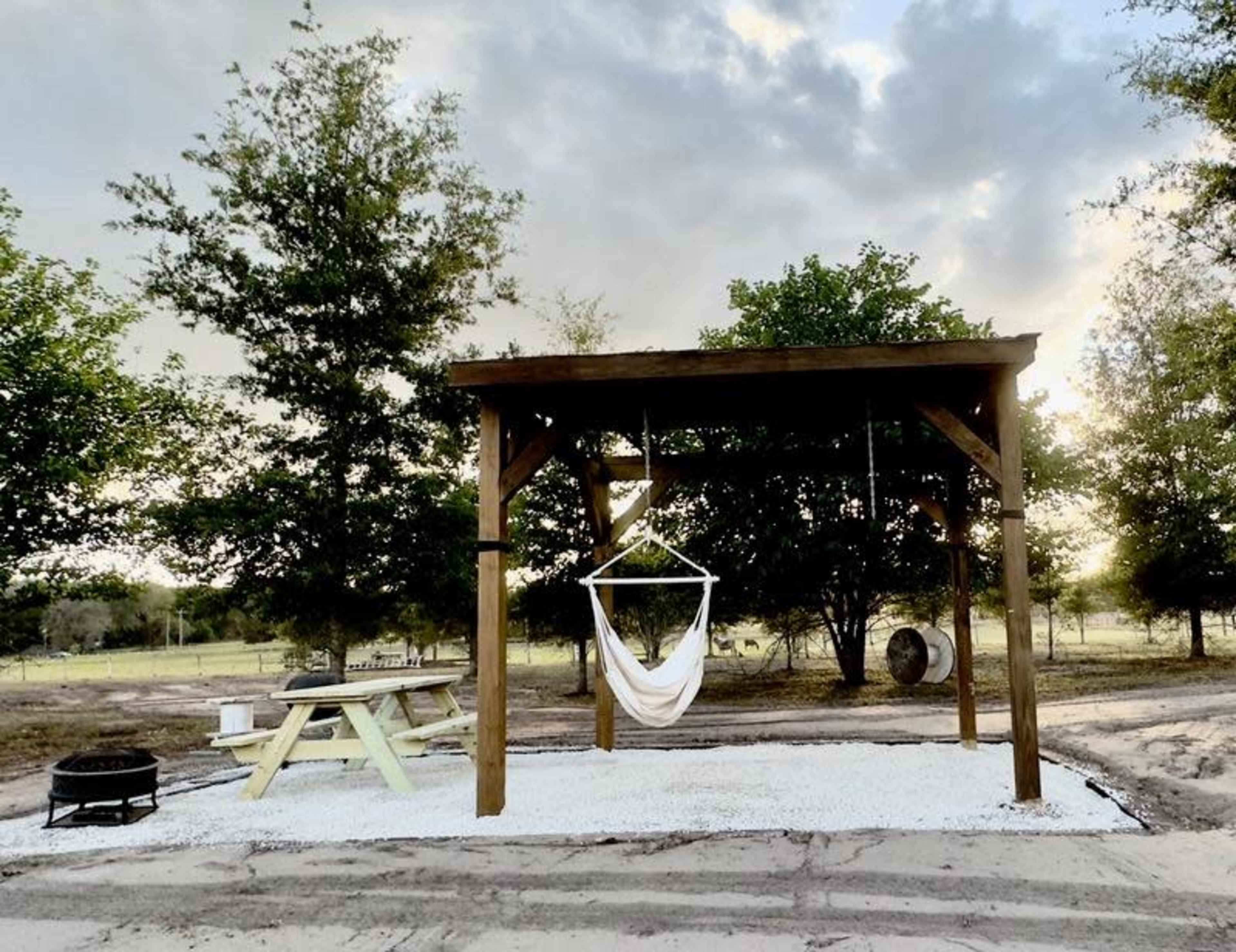 A wooden structure with a hammock, picnic table, and fire pit set on a gravel area surrounded by trees and a cloudy sky.