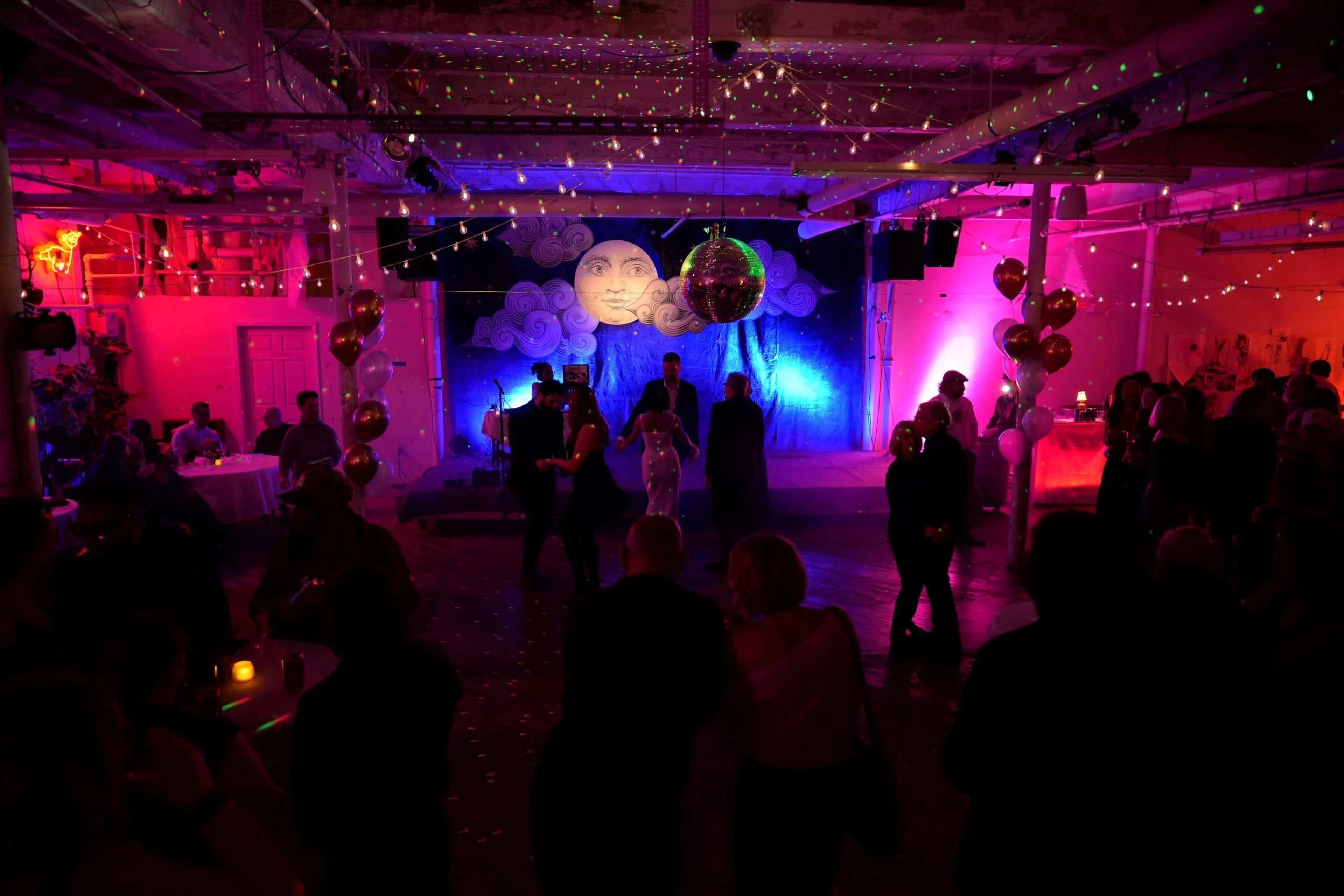 A dimly lit venue hosts a lively gathering with colorful lighting, a large mural of a moon, and people dancing amid decorations and balloons.