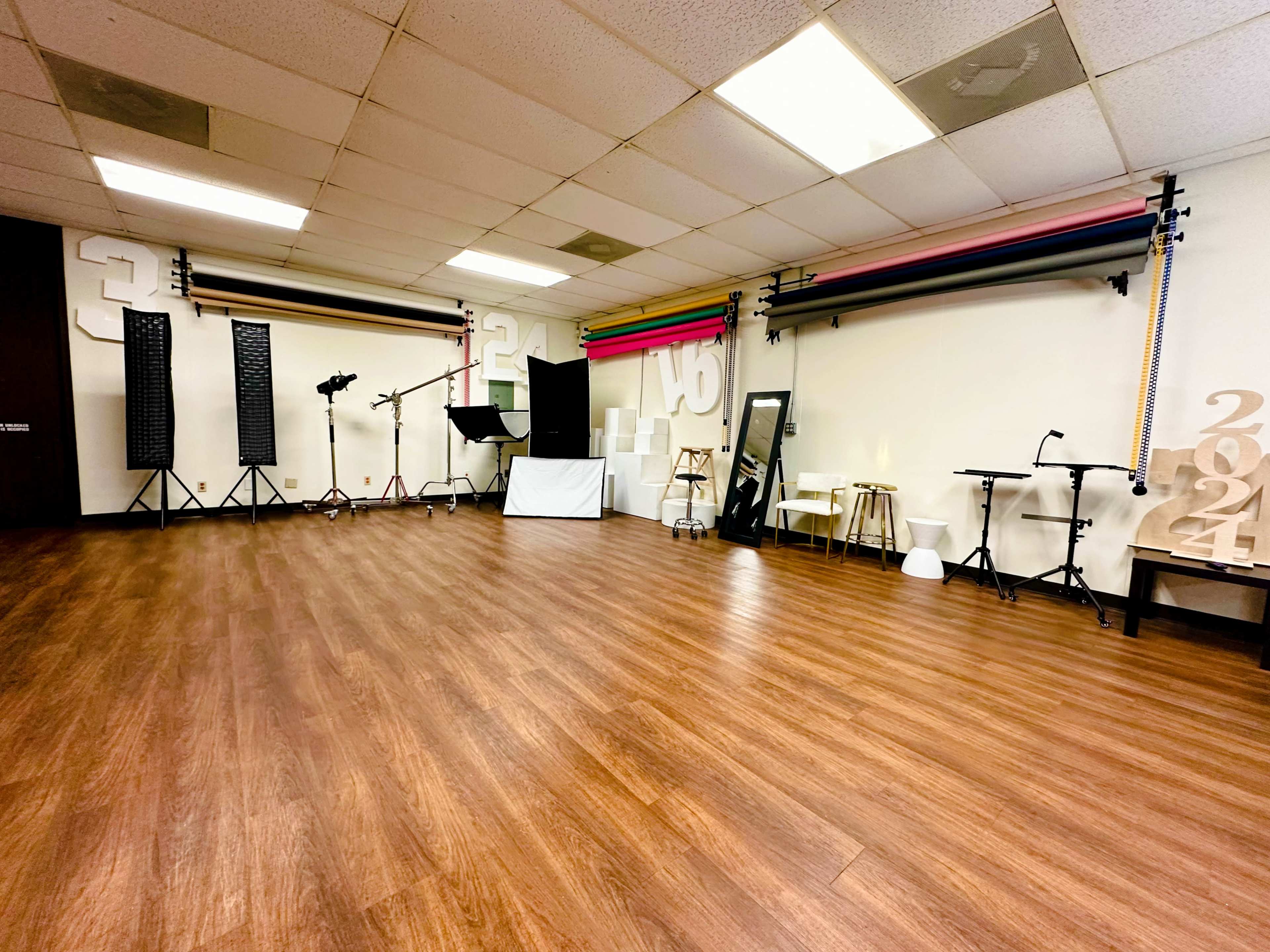 A spacious studio with wooden flooring, equipped with various backdrop rolls, lighting stands, and a mirror.
