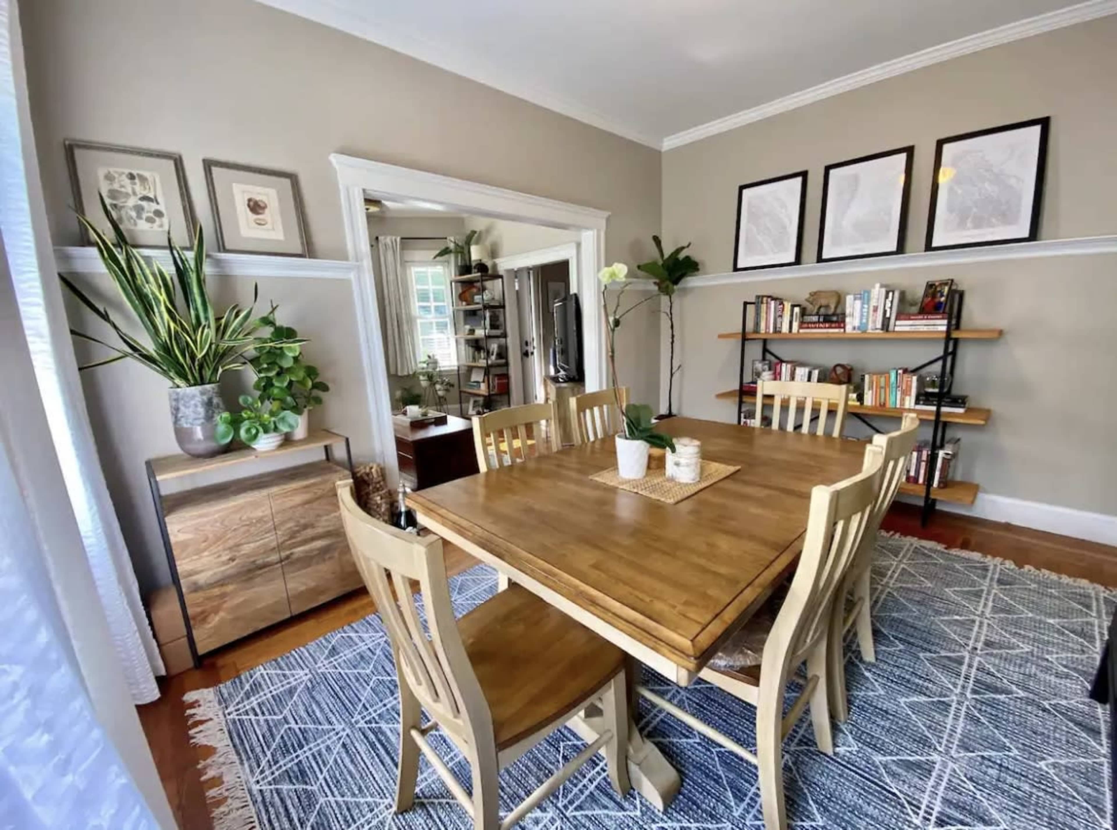 A dining room features a wooden table surrounded by four chairs, with plants and bookshelves in the background, and framed pictures on the wall.