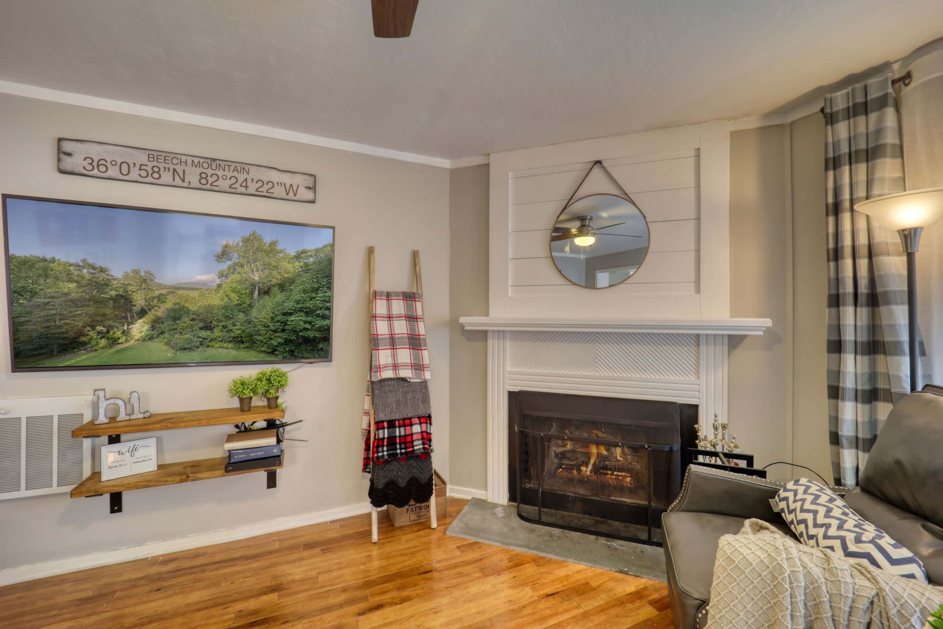 A living room features a large wall-mounted television displaying a landscape, with a fireplace, a decorative mirror above the mantel, and a wooden shelf holding a plant and decorative items.