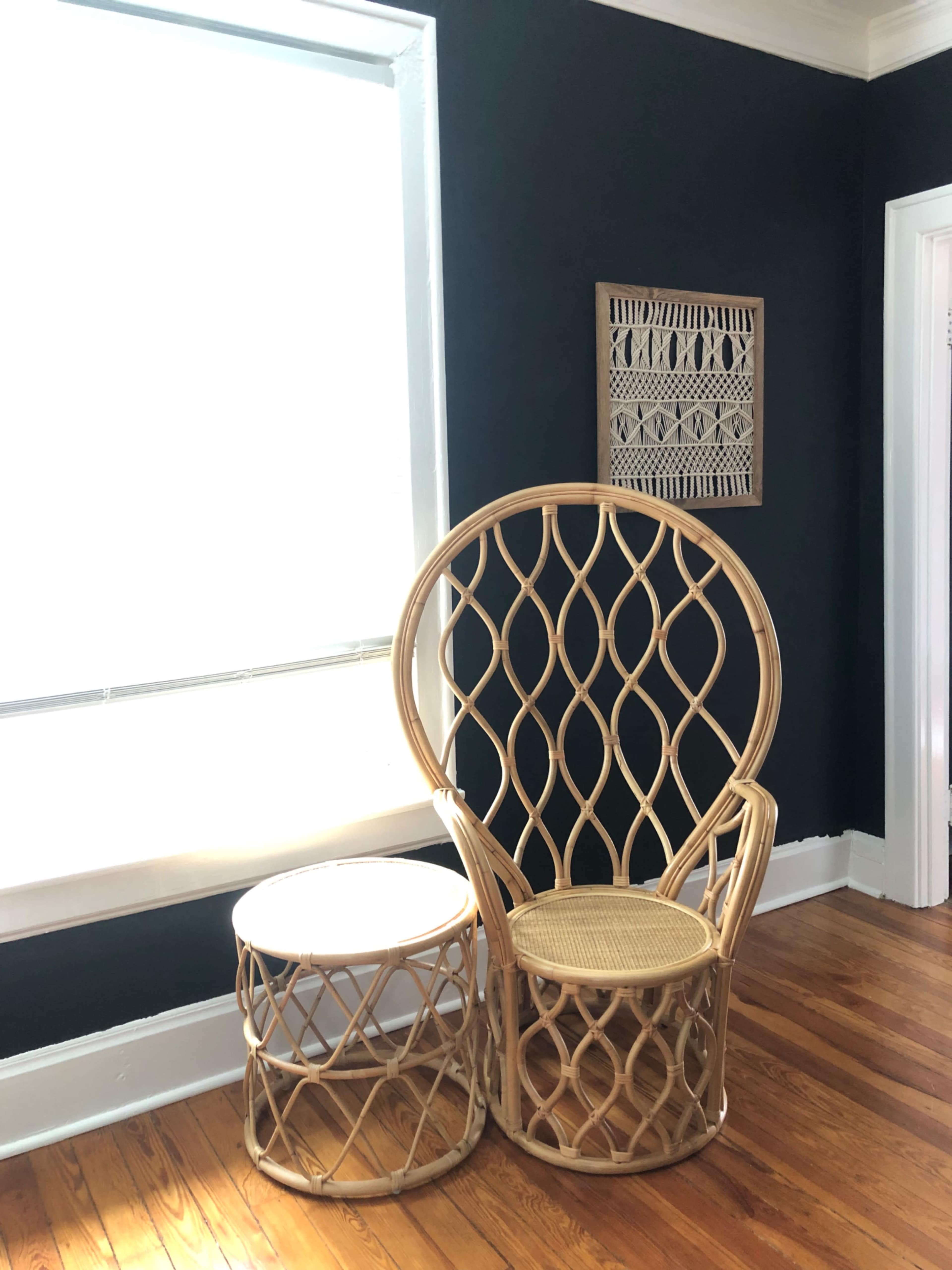 A large rattan chair with an intricate design and a round side table are positioned beside a window in a room with dark walls.