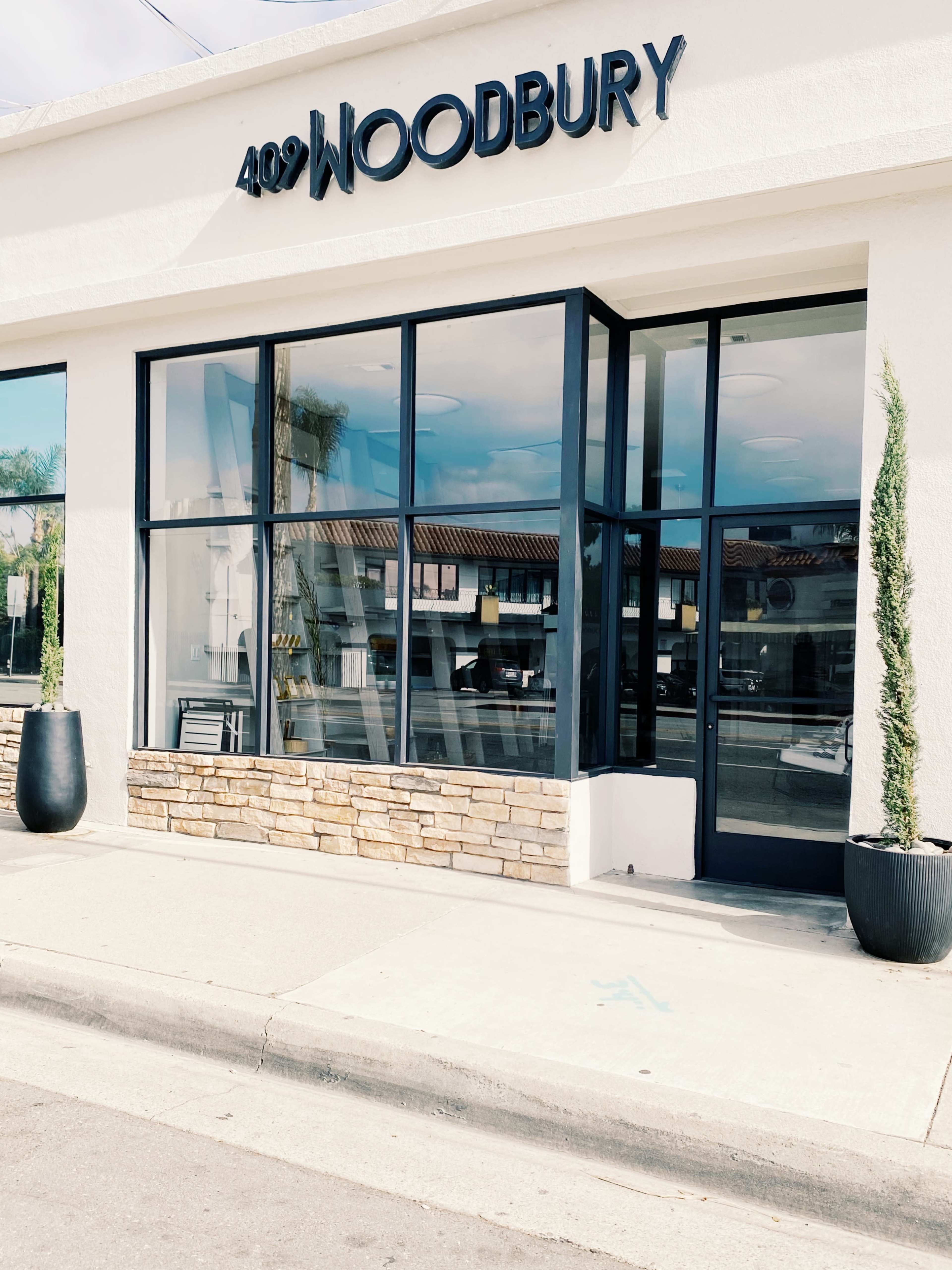 The exterior of a building with the address "402 WOODBURY" displayed above large windows and planters on either side of the entrance.