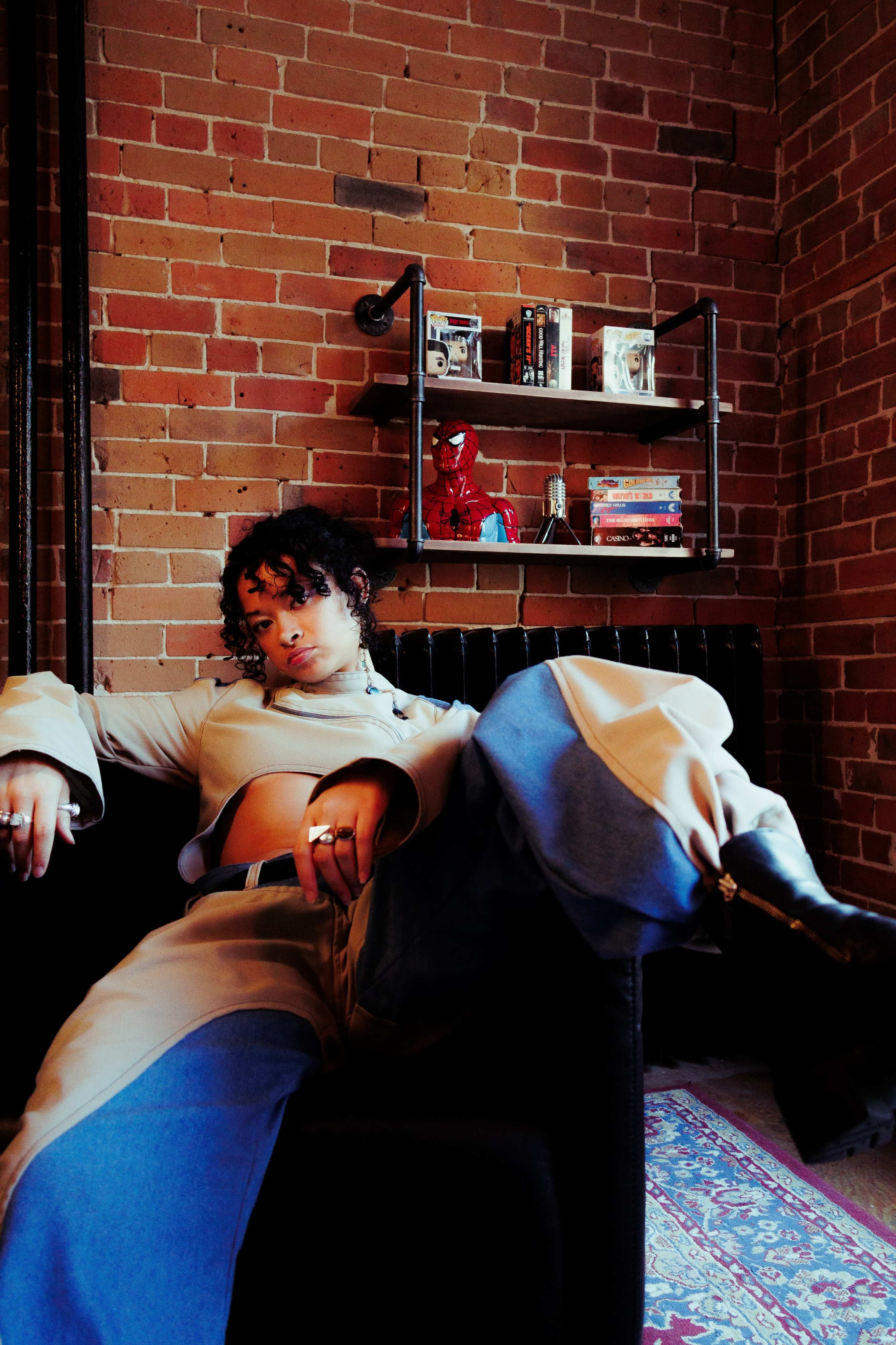 A person with curly hair sits casually on a couch in a brick-walled room, surrounded by shelves filled with books and a Spider-Man figure.