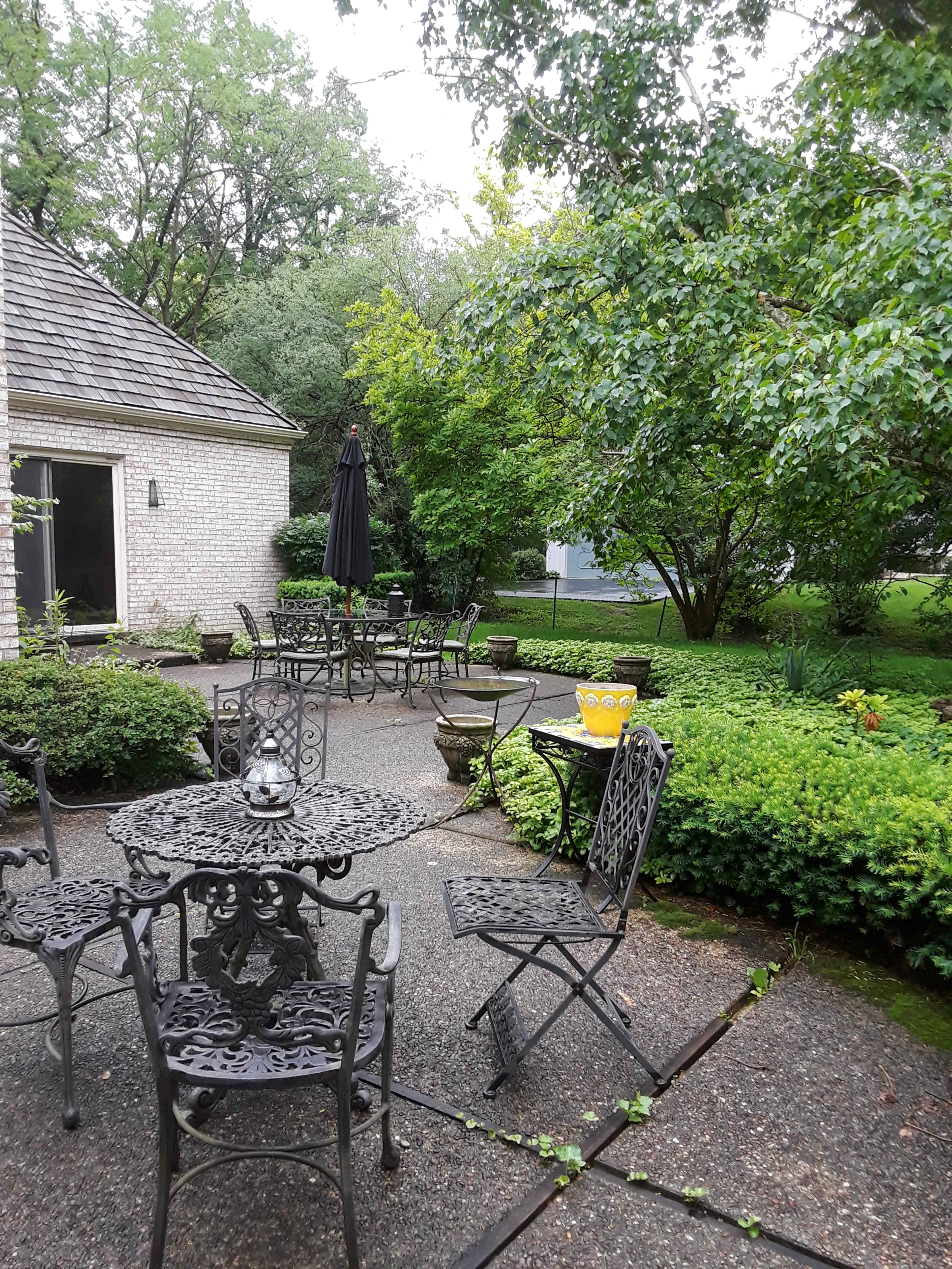 A courtyard features metal tables and chairs surrounded by lush greenery and a stone pathway.