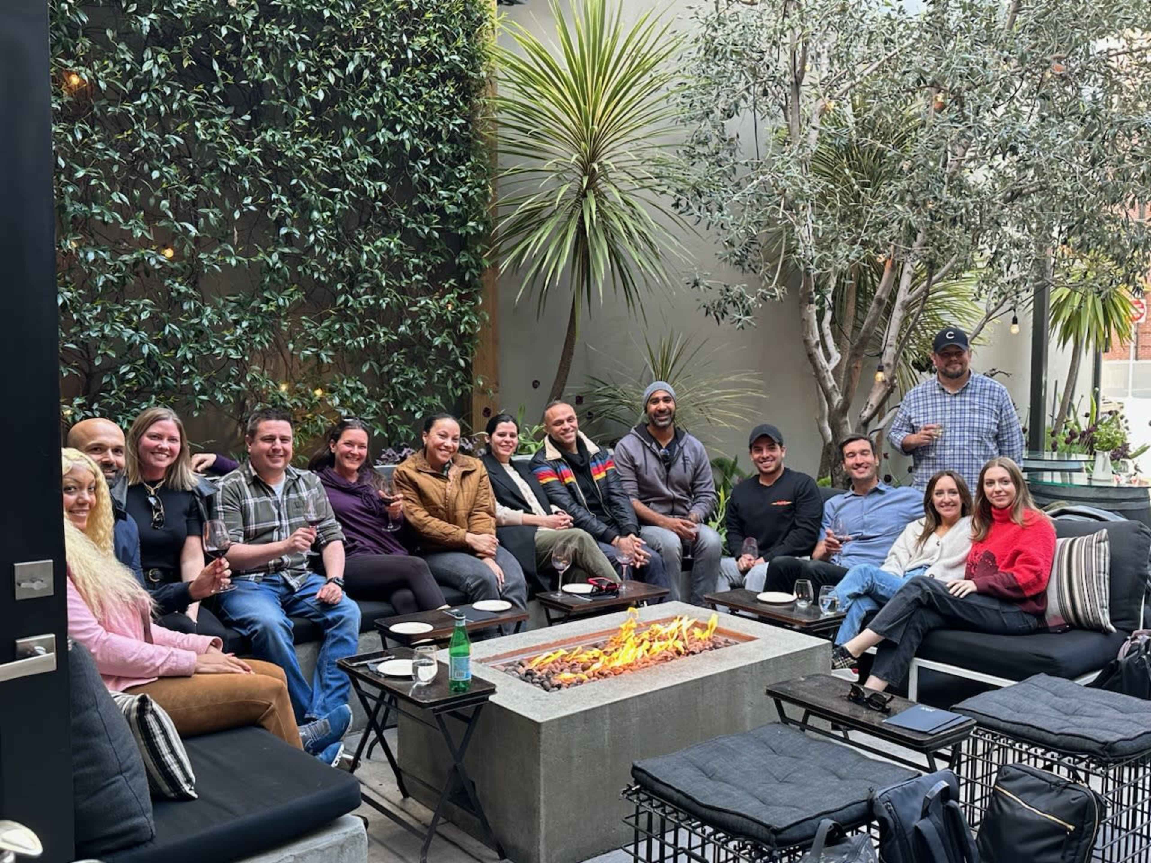 A group of people gathers around a fire pit in a lush outdoor seating area.
