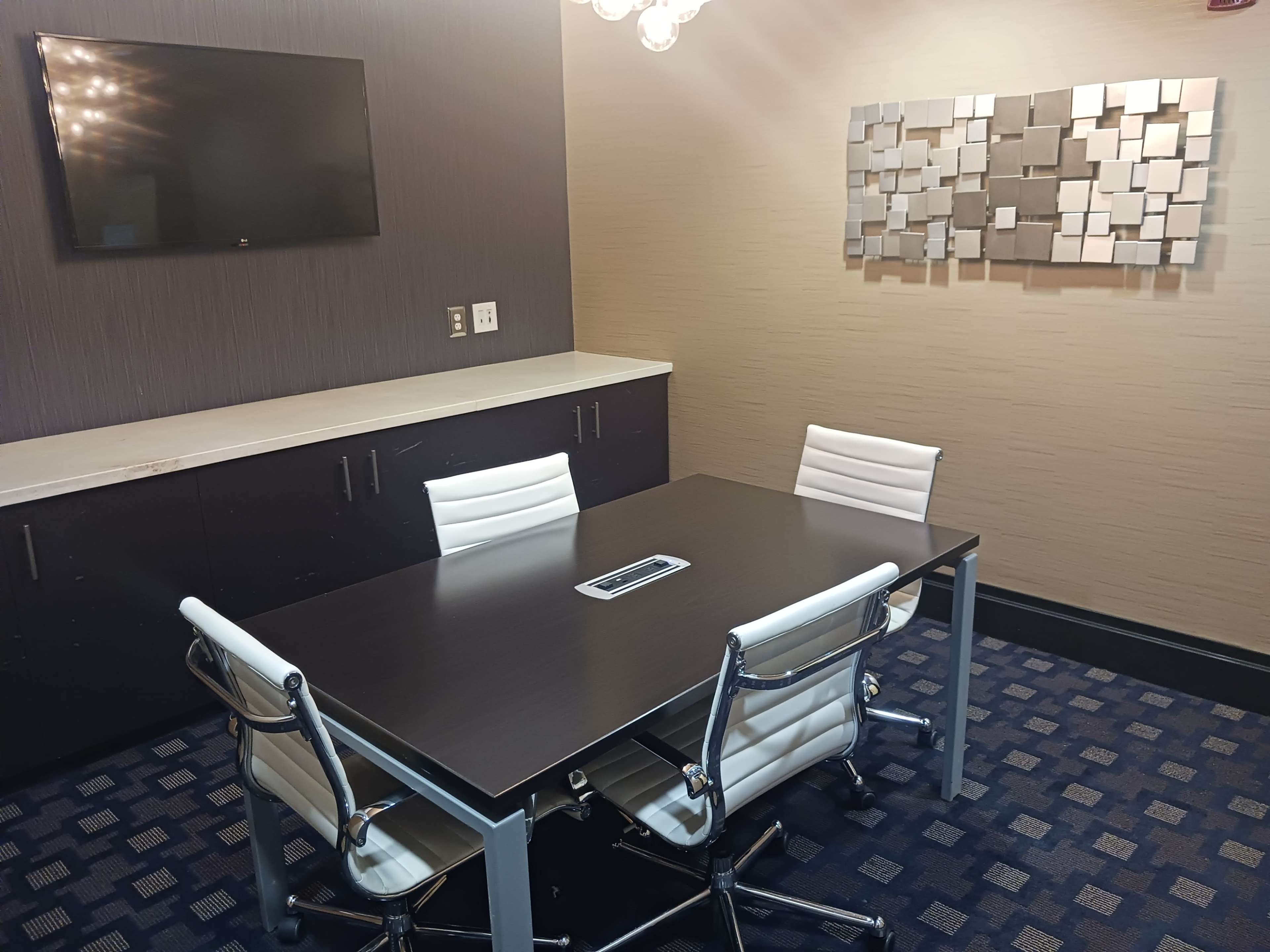A modern conference room with a rectangular table surrounded by four white chairs, a wall-mounted screen, and abstract wall art.