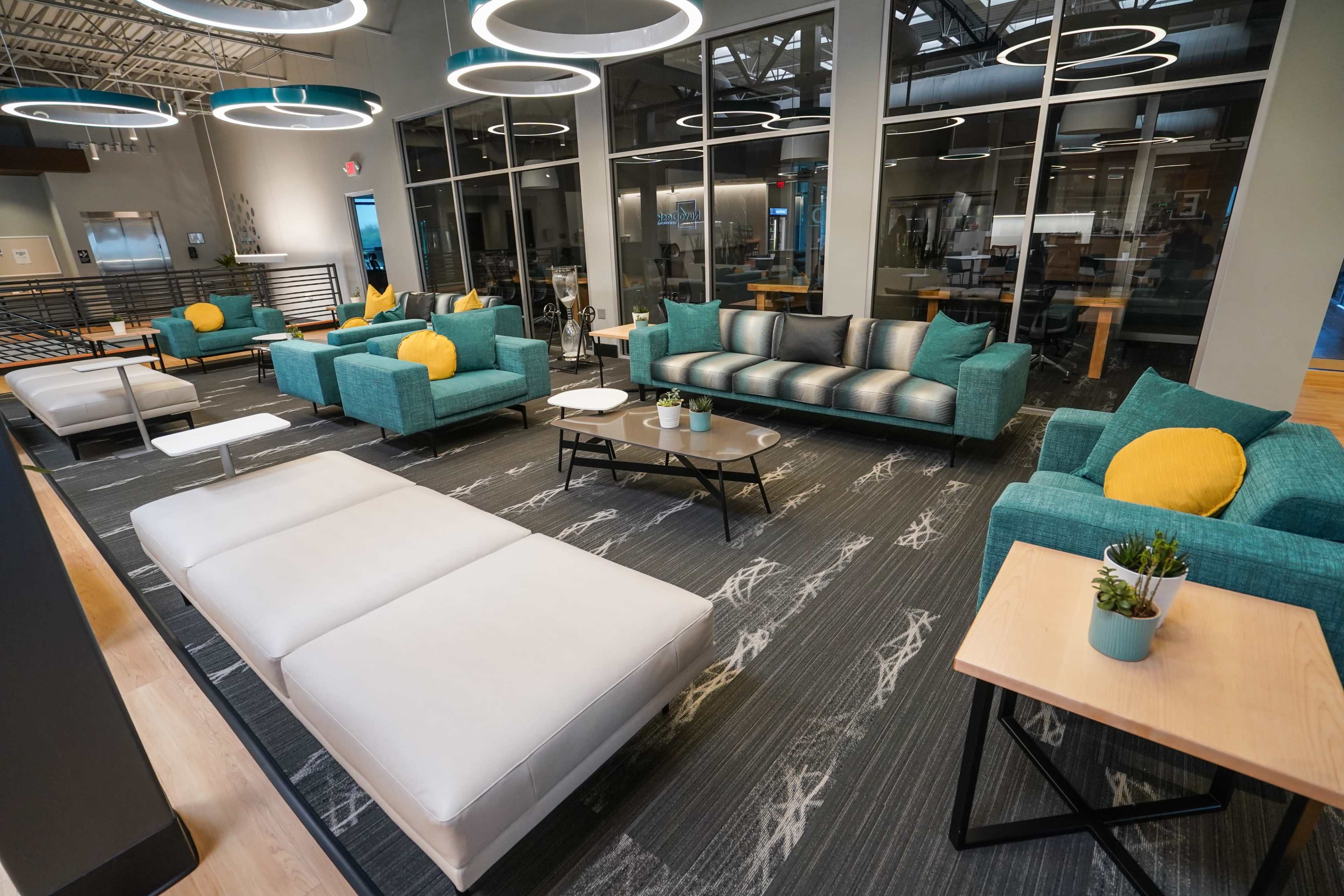 The image shows a modern lounge area with teal and gray seating, a white sectional sofa, and a coffee table on a patterned rug, surrounded by large windows.