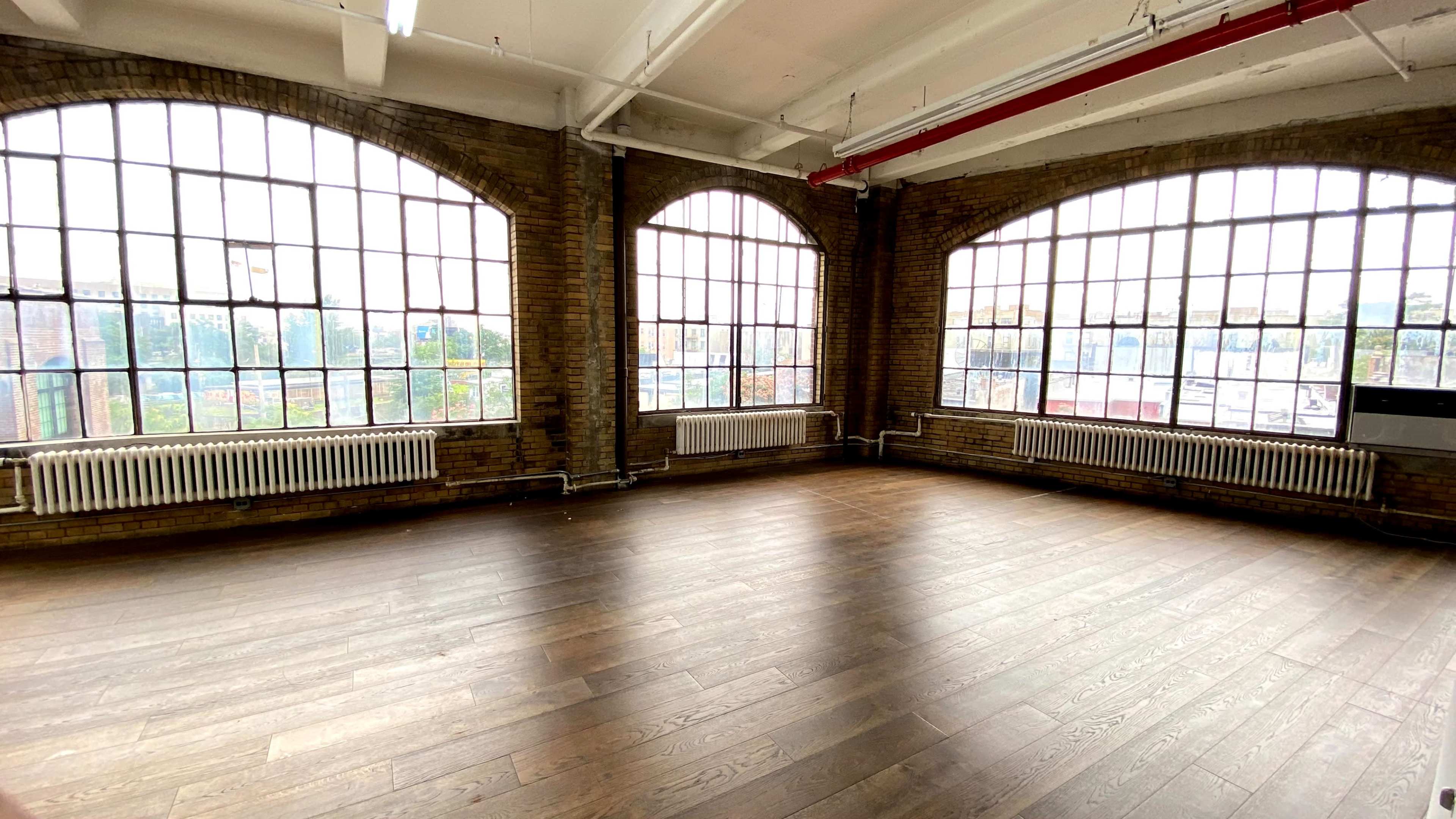 A spacious room with largearched windows and wooden floors, featuring exposed brick walls and visible heating radiators.