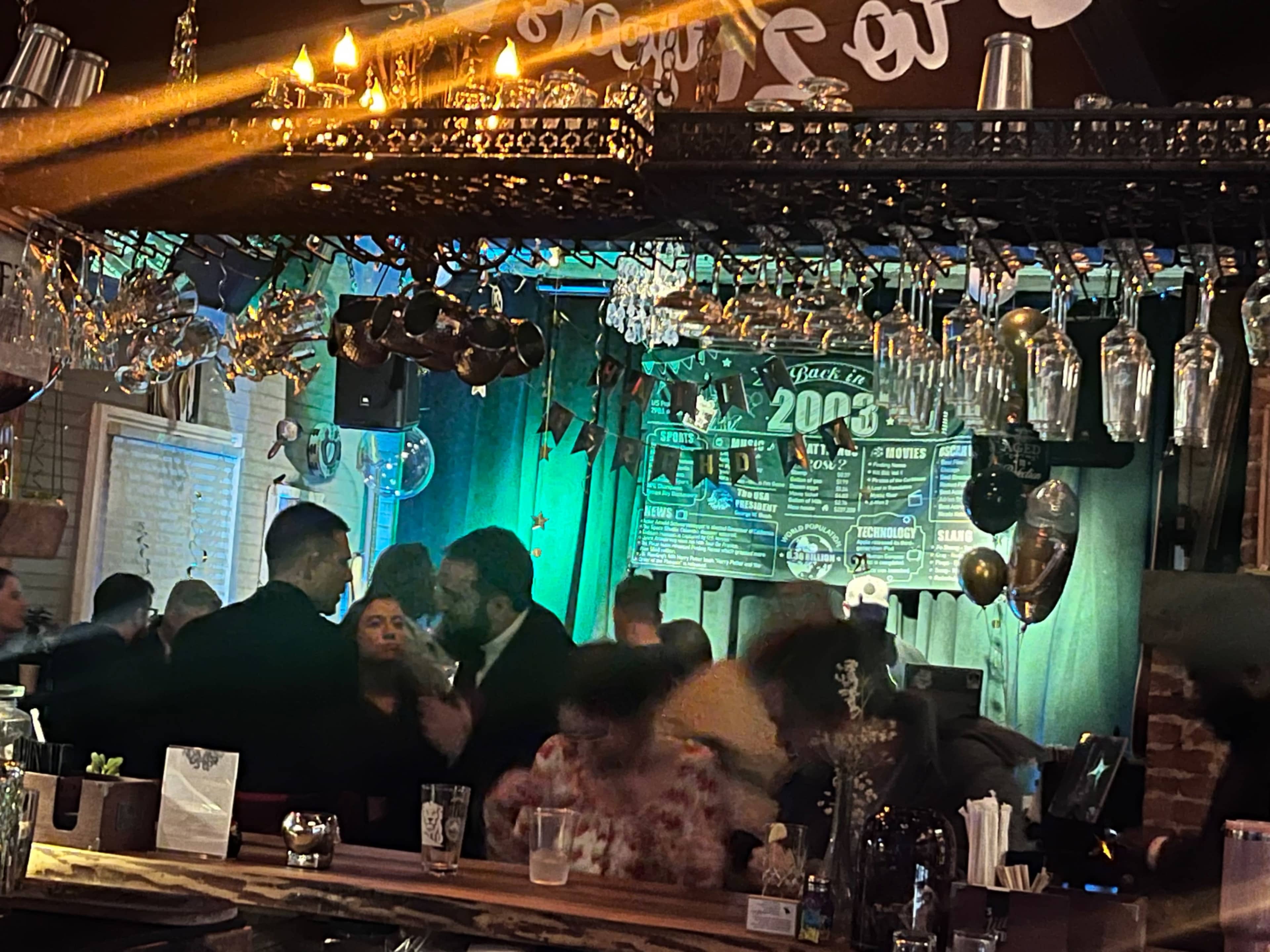 A vibrant bar scene shows people socializing under dim lighting, with decorative glassware hanging above and a promotional poster on the wall.