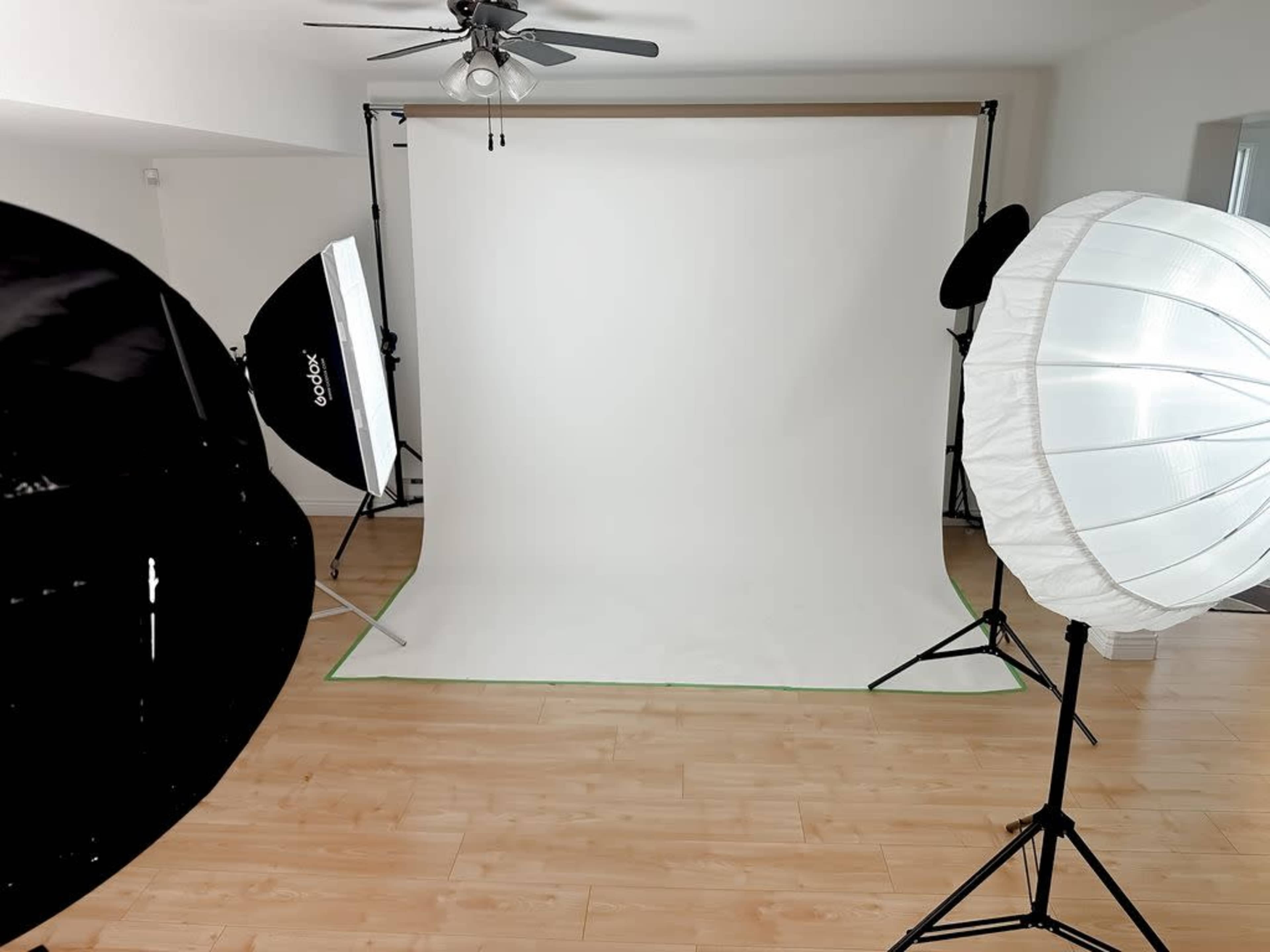 The image shows a photography studio setup featuring a white backdrop, several softbox lights, and wooden flooring.